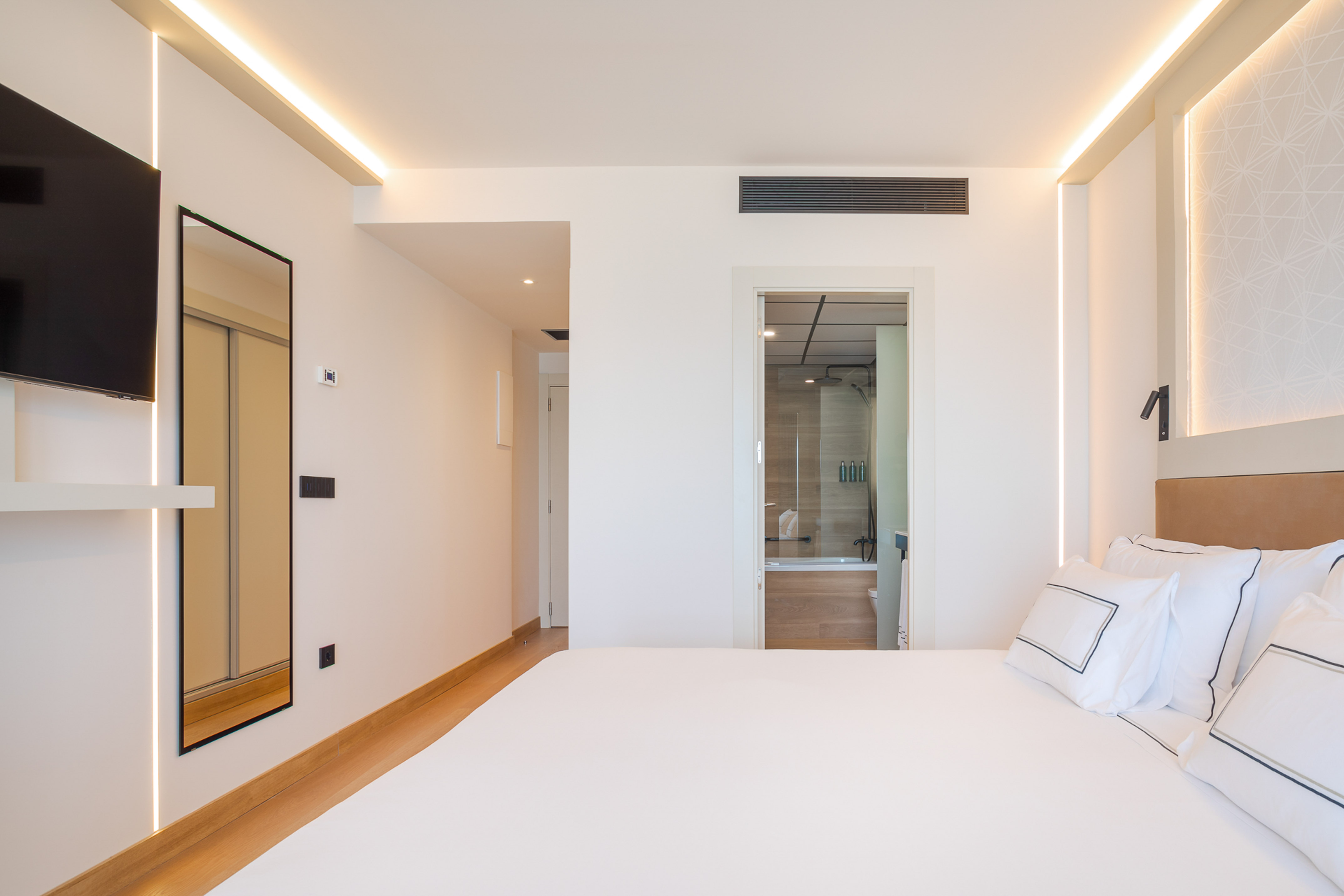 Modern hotel bedroom with white bedding, TV, LED lighting, and ensuite bathroom.