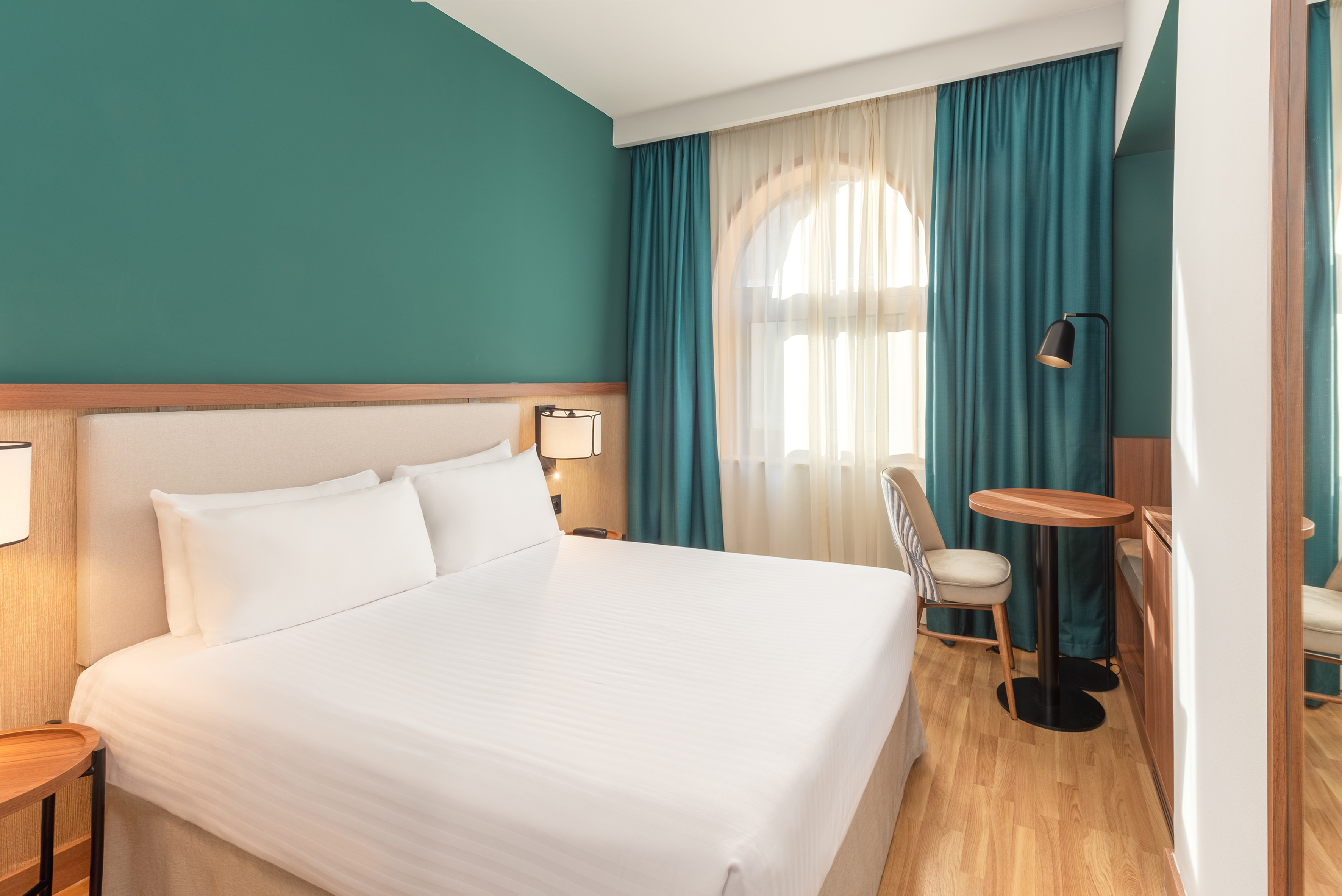 Hotel room with white bed, teal walls, arched window, and wooden table.