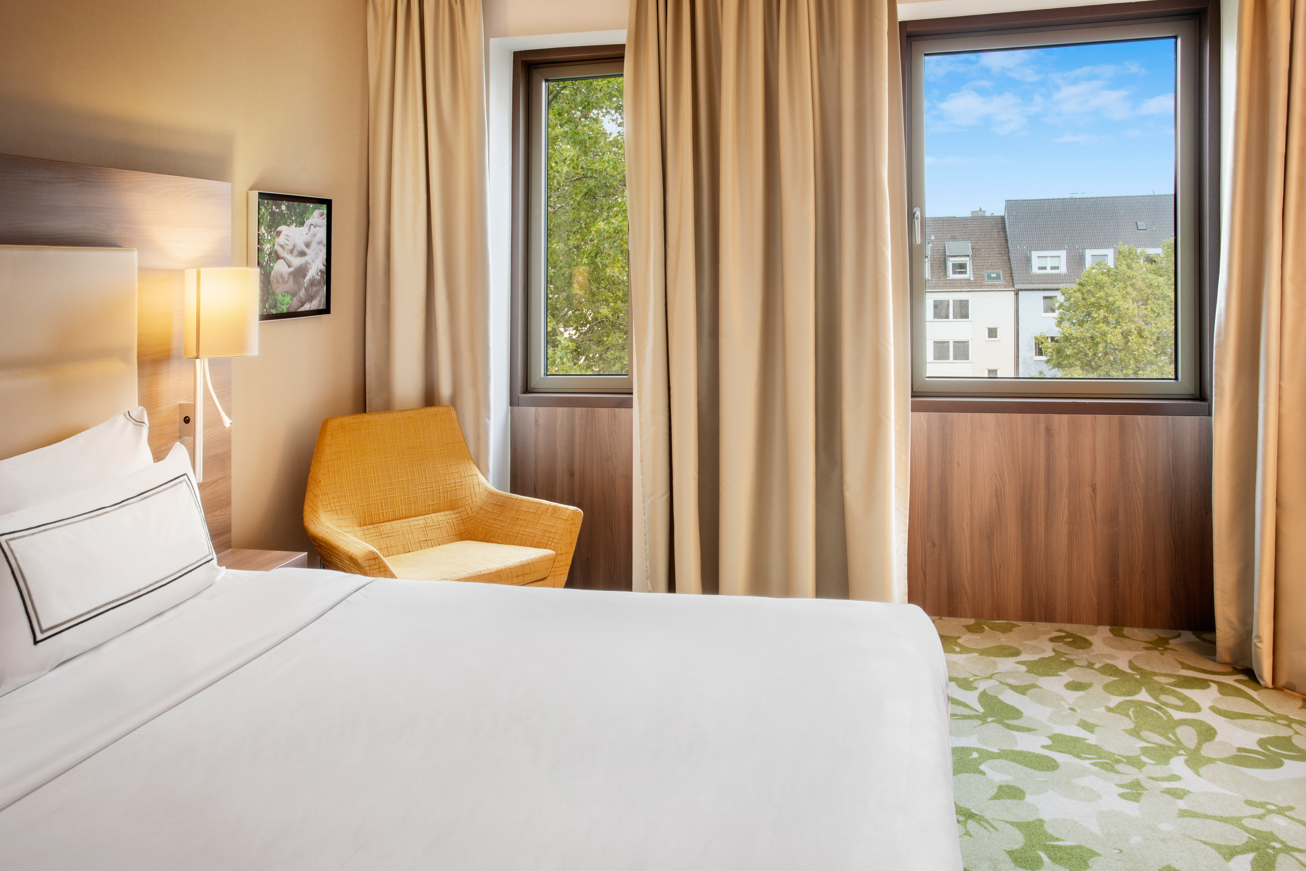 Modern hotel bedroom with white bed, yellow armchair, windows, and city view.
