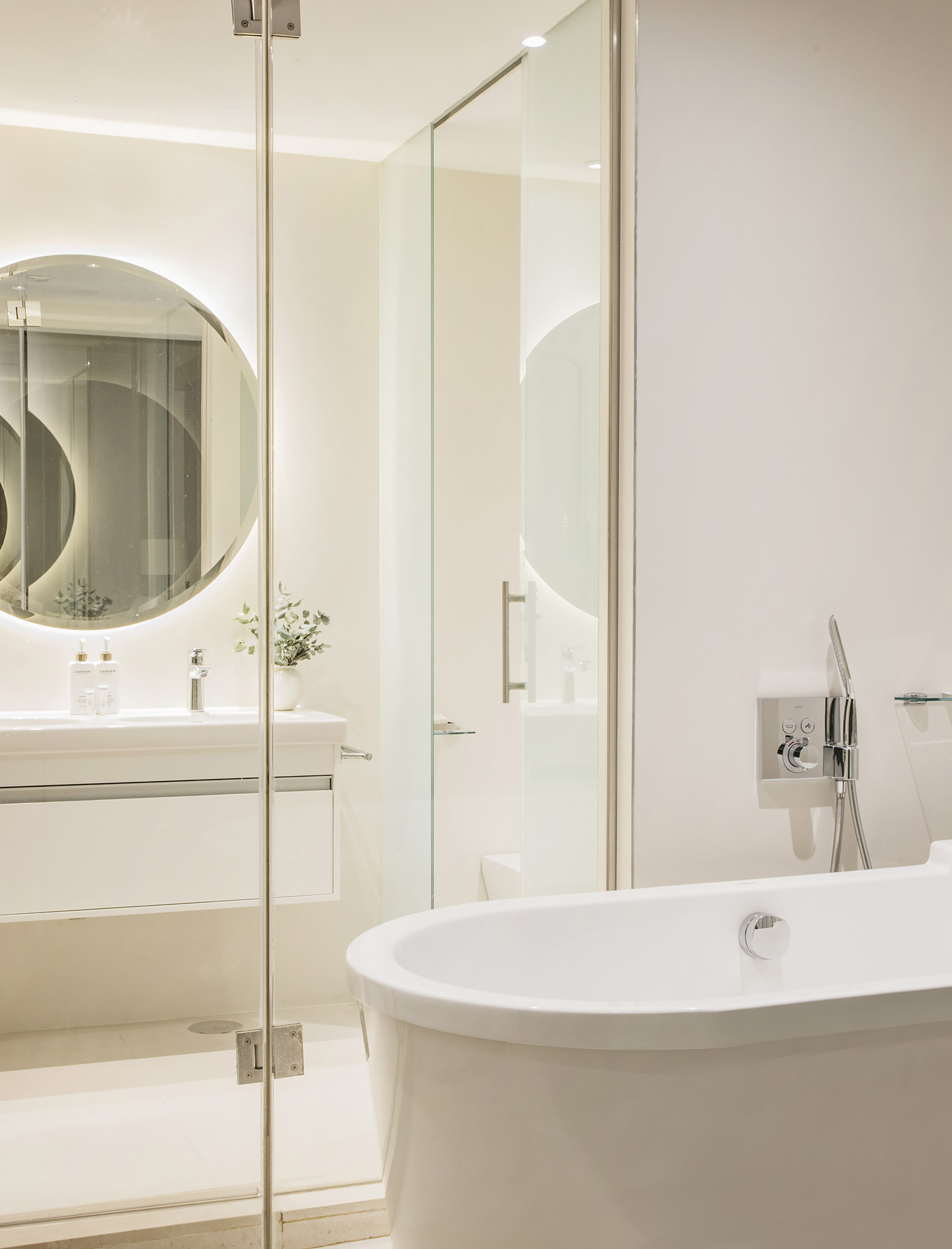 Modern white bathroom with freestanding tub, glass shower, backlit vanity mirror.