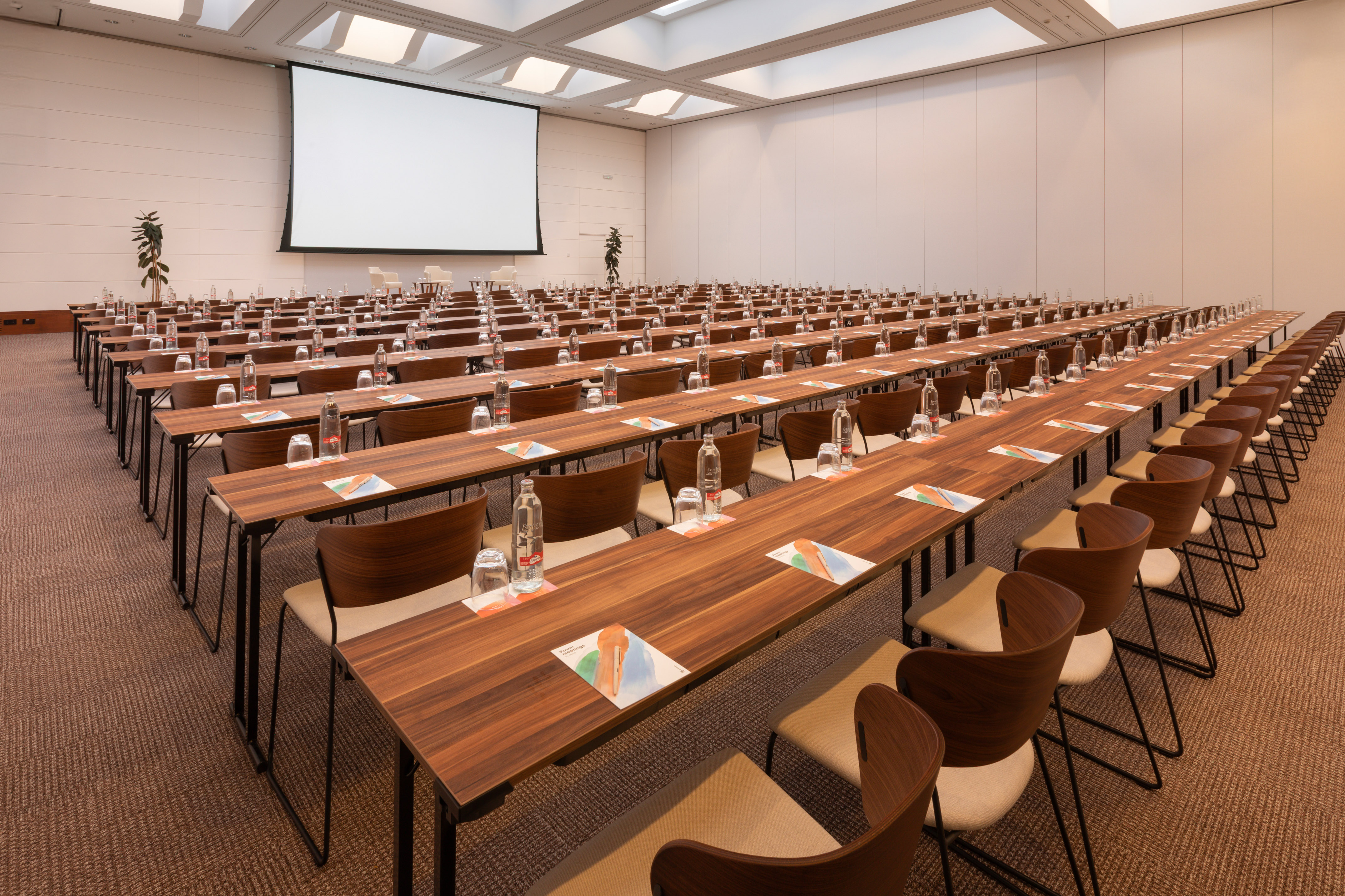 Conference room with rows of tables, chairs, water, and a large projection screen.