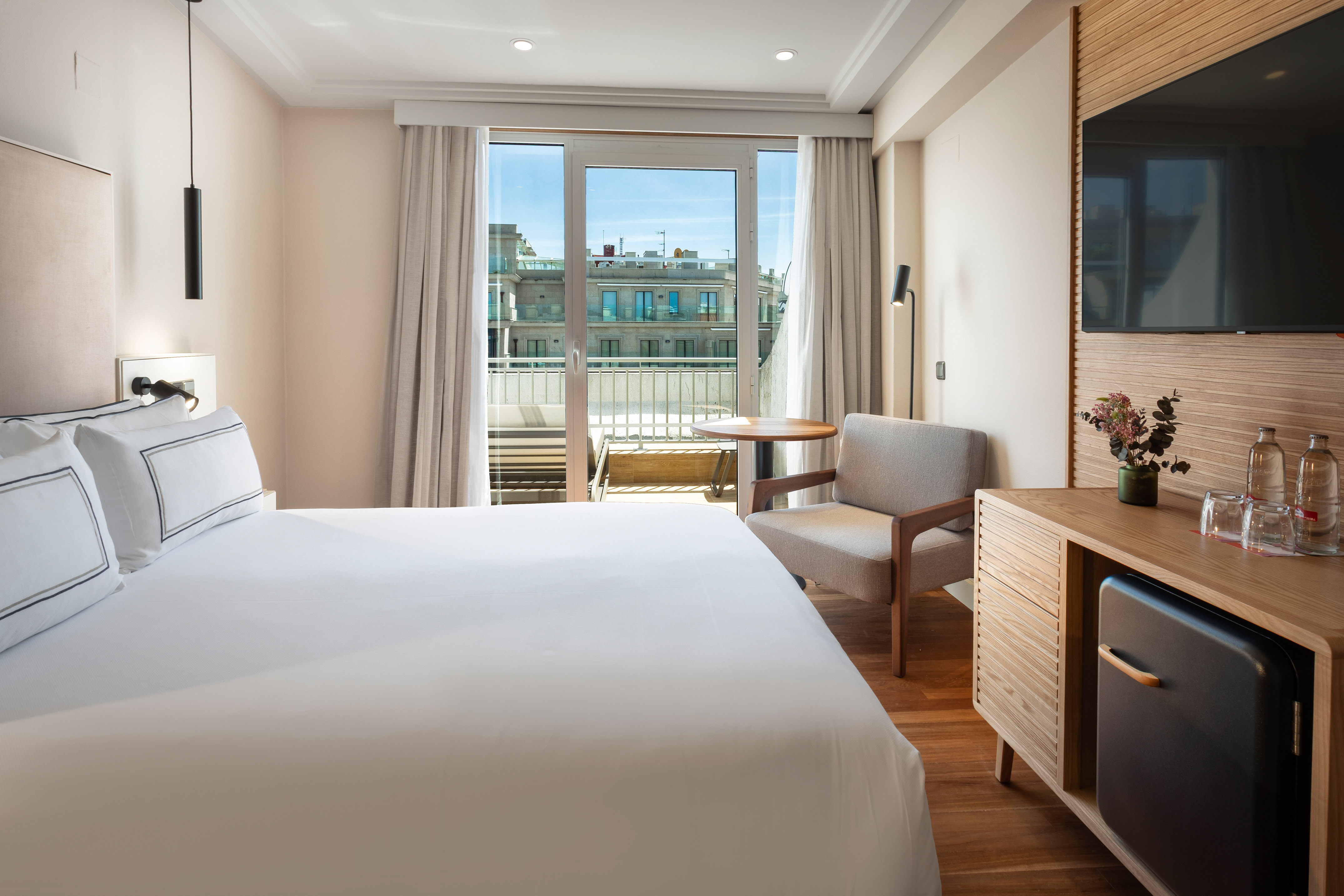 Modern hotel room with large bed, city view balcony, TV, and wooden mini-bar.