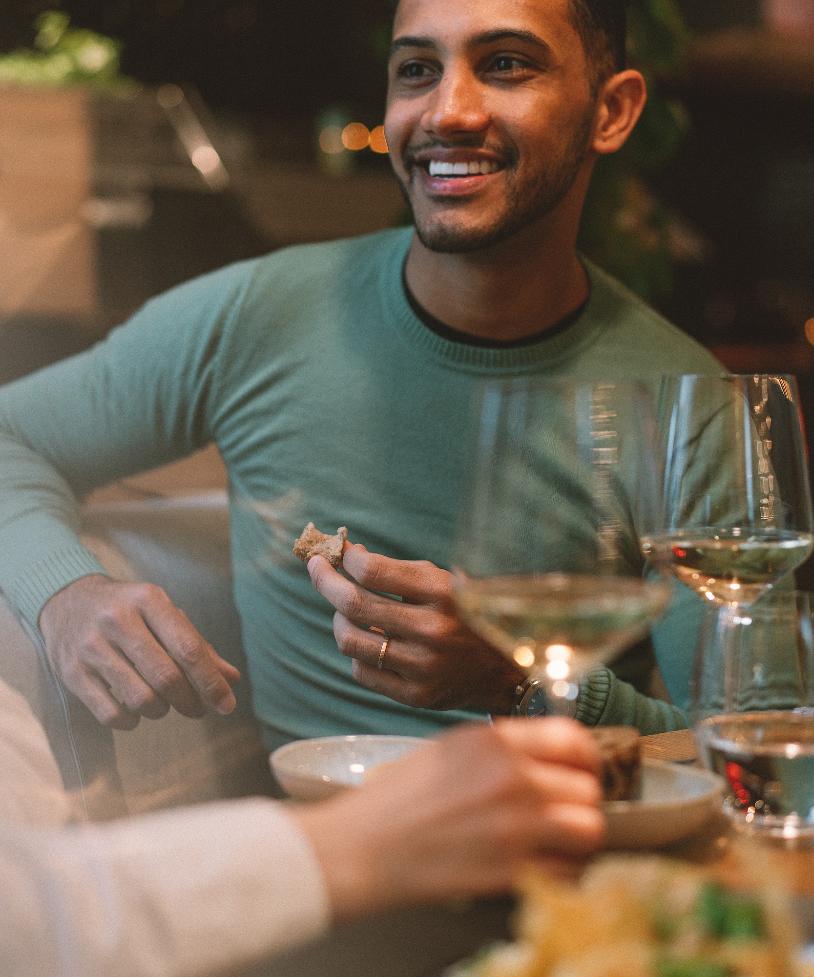 Smiling man in green sweater holding bread, dining with wine.