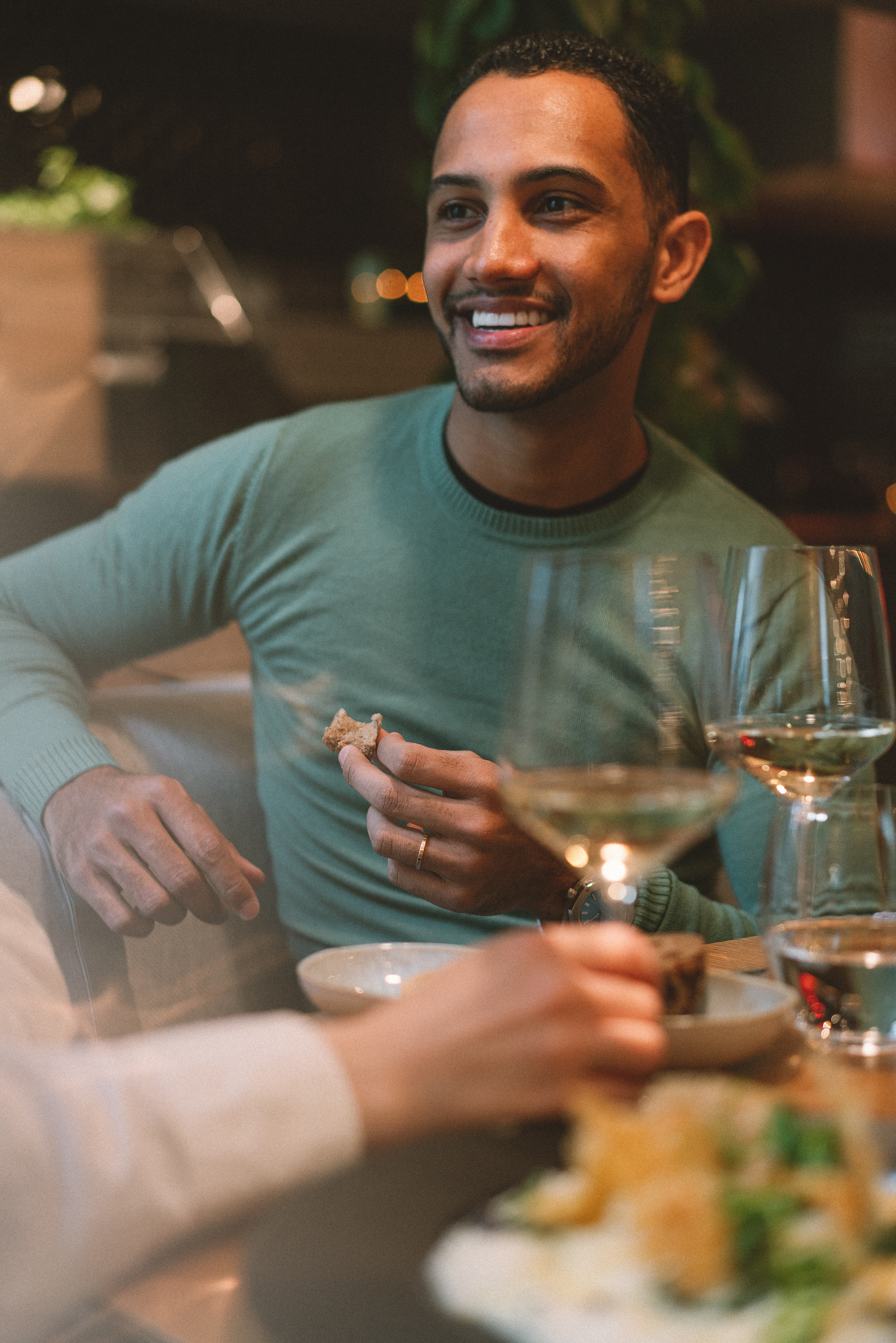 Smiling man in green sweater holding bread, dining with wine.