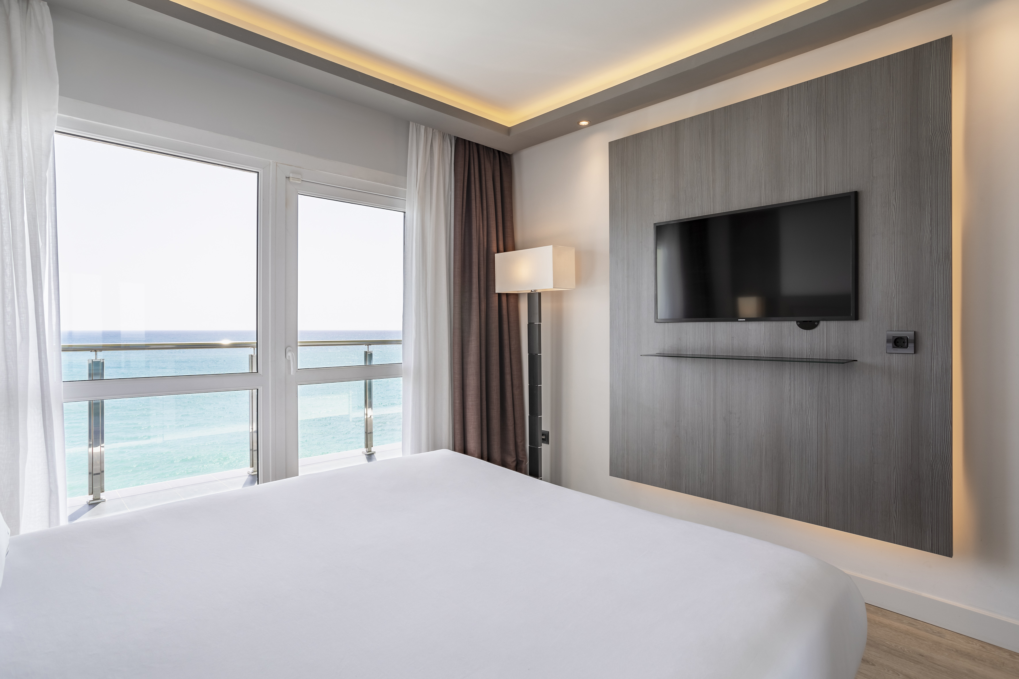 Modern hotel bedroom with ocean view, white bed, wall TV, and accent lighting.