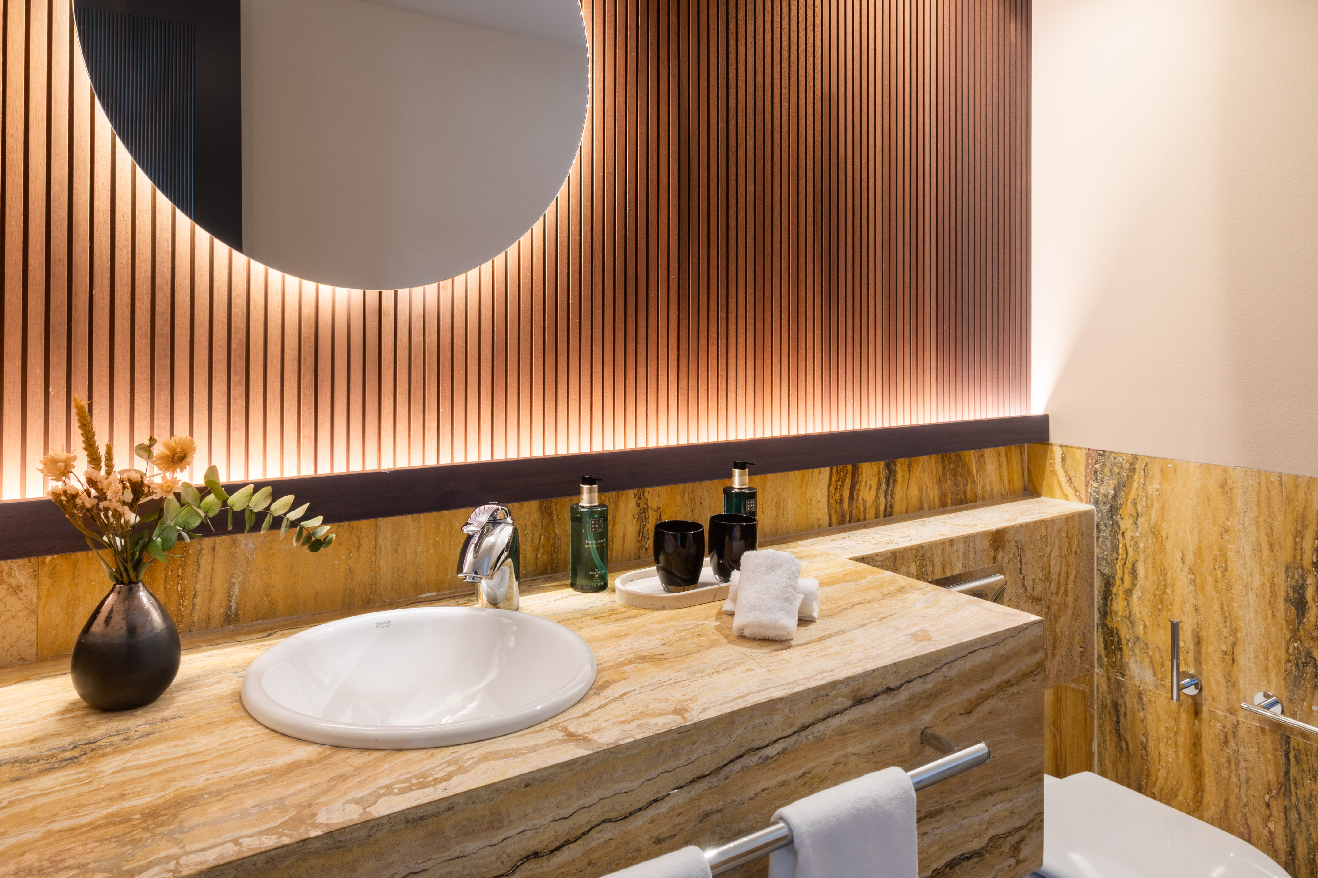 Modern bathroom with marble vanity, circular backlit mirror, and wood slat wall.