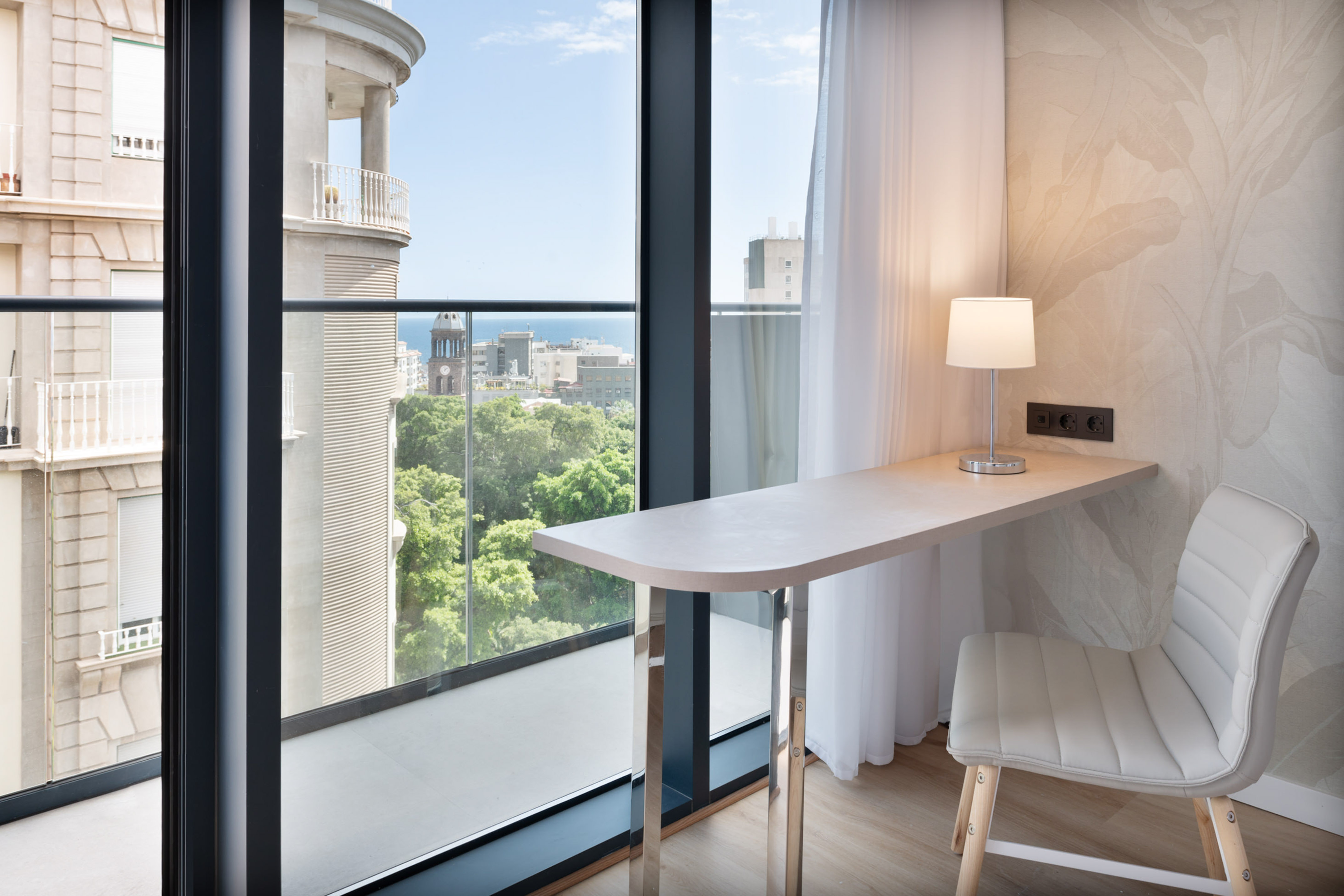 Hotel room workspace with desk, lamp, and large window overlooking city and sea.