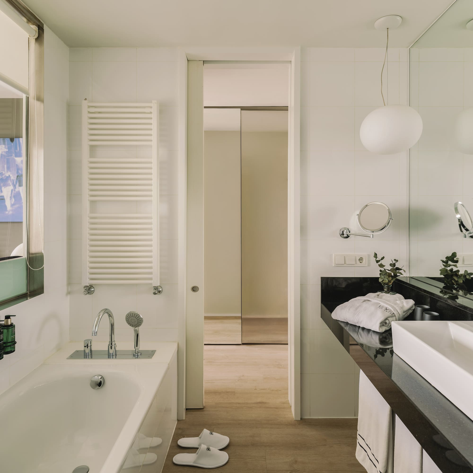 Modern hotel bathroom with white bathtub, sink, black counter, and wood floor.