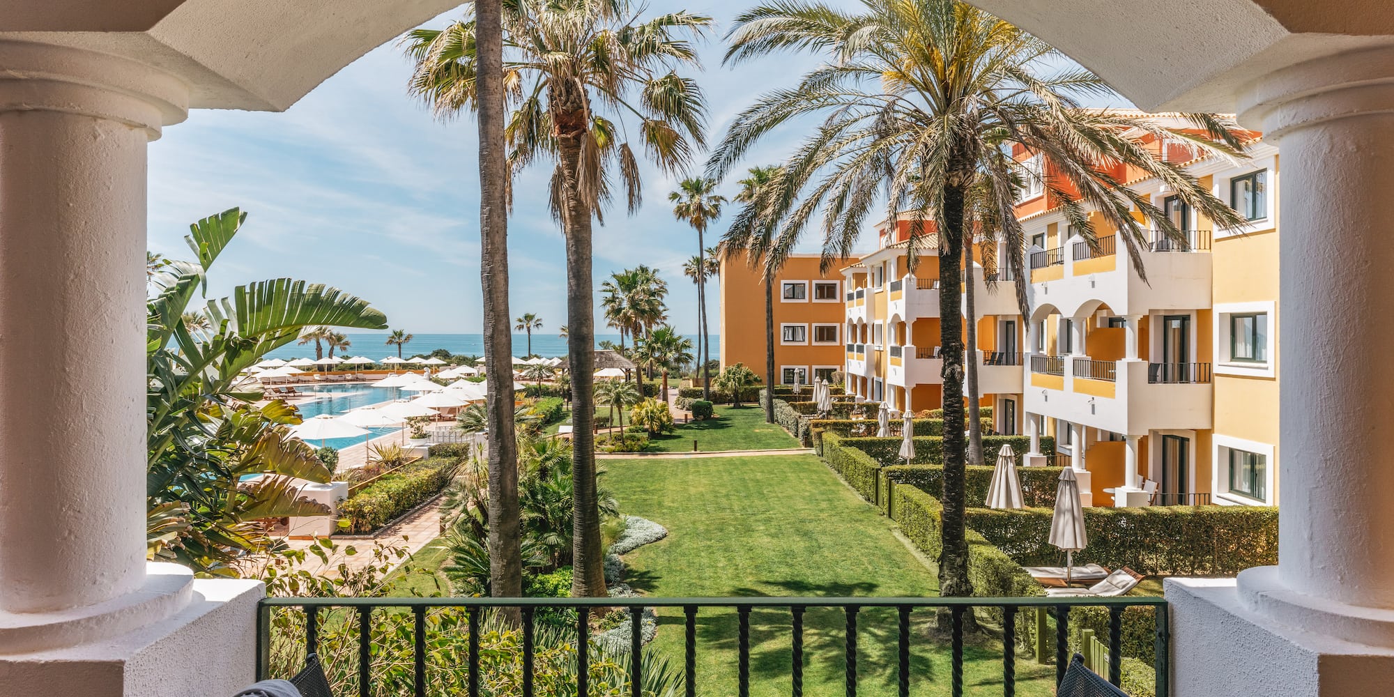 Tropical resort balcony view: ocean, pool, palm trees, hotel buildings.