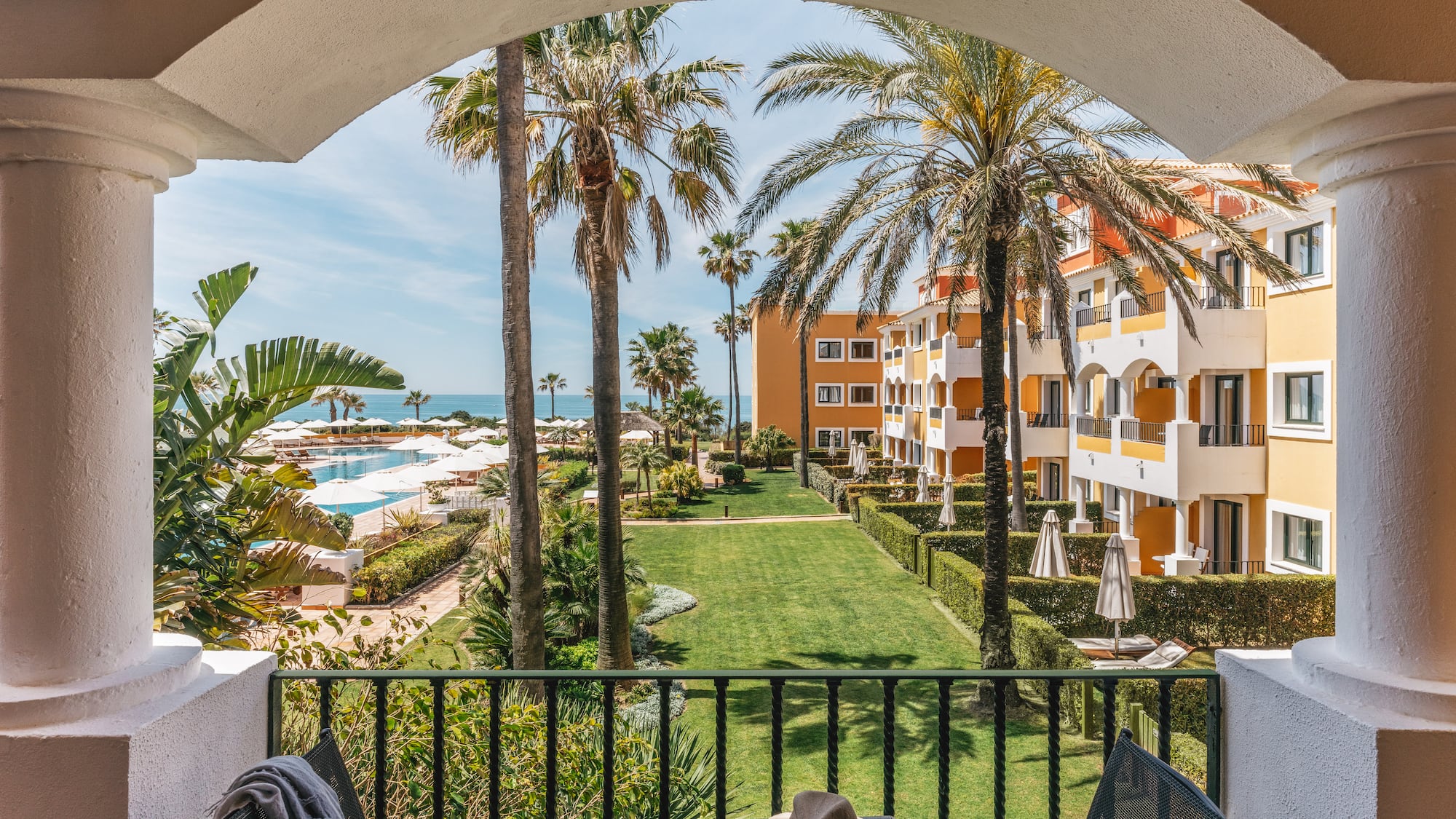 Tropical resort balcony view: ocean, pool, palm trees, hotel buildings.