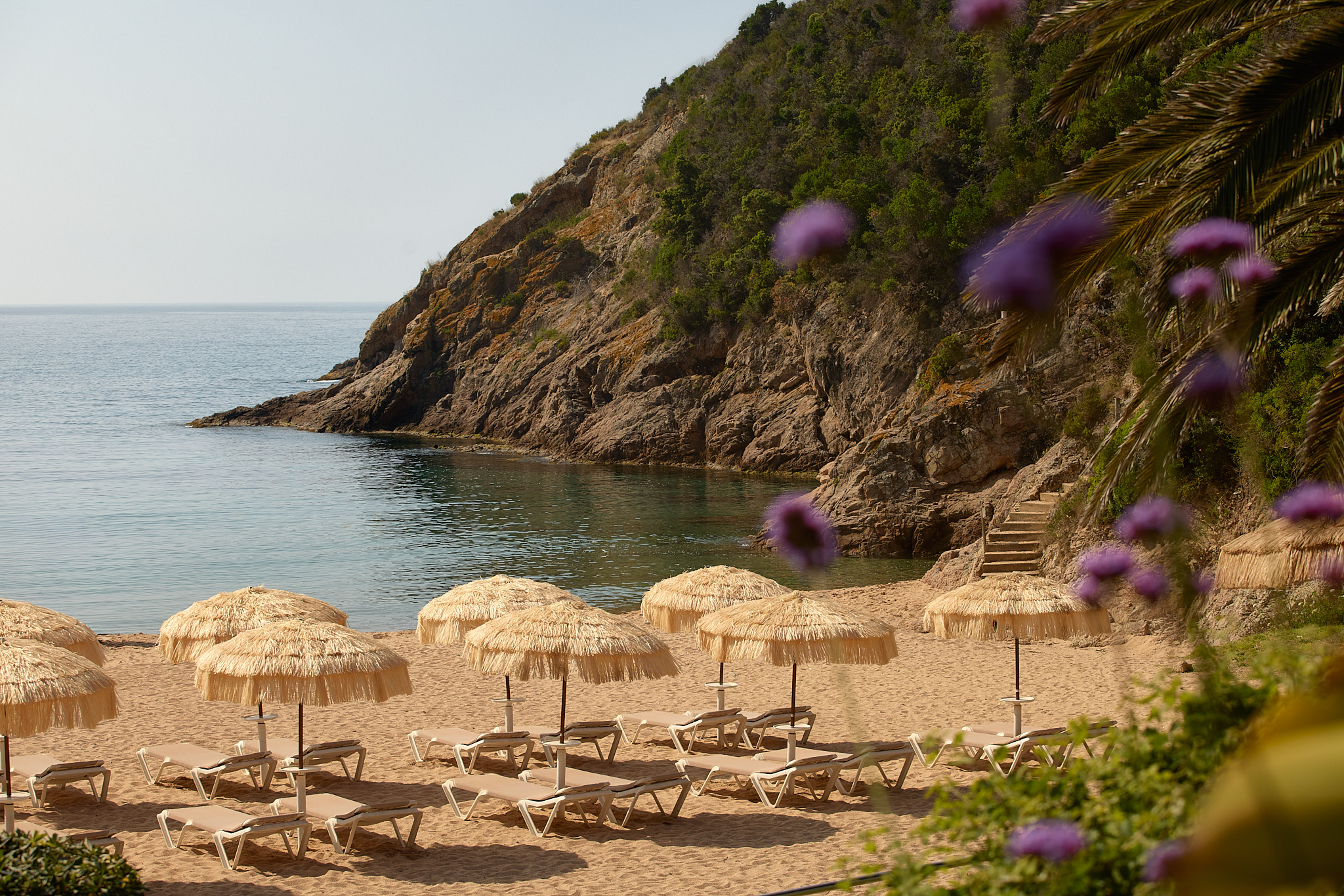 Sandy beach, straw umbrellas, loungers, calm sea, and rocky cliff.