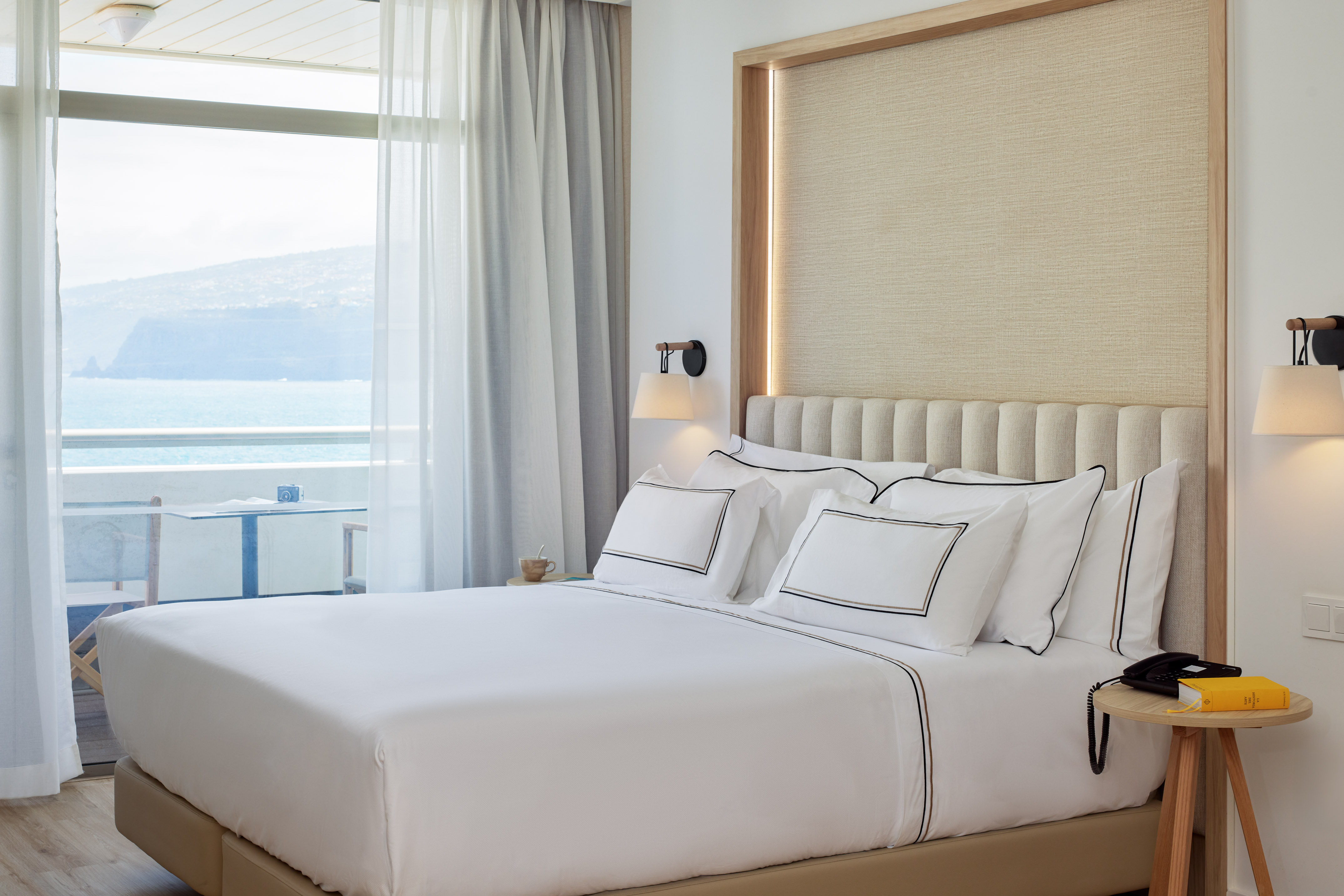 Modern hotel room with sea view balcony, white bed, and modern decor.