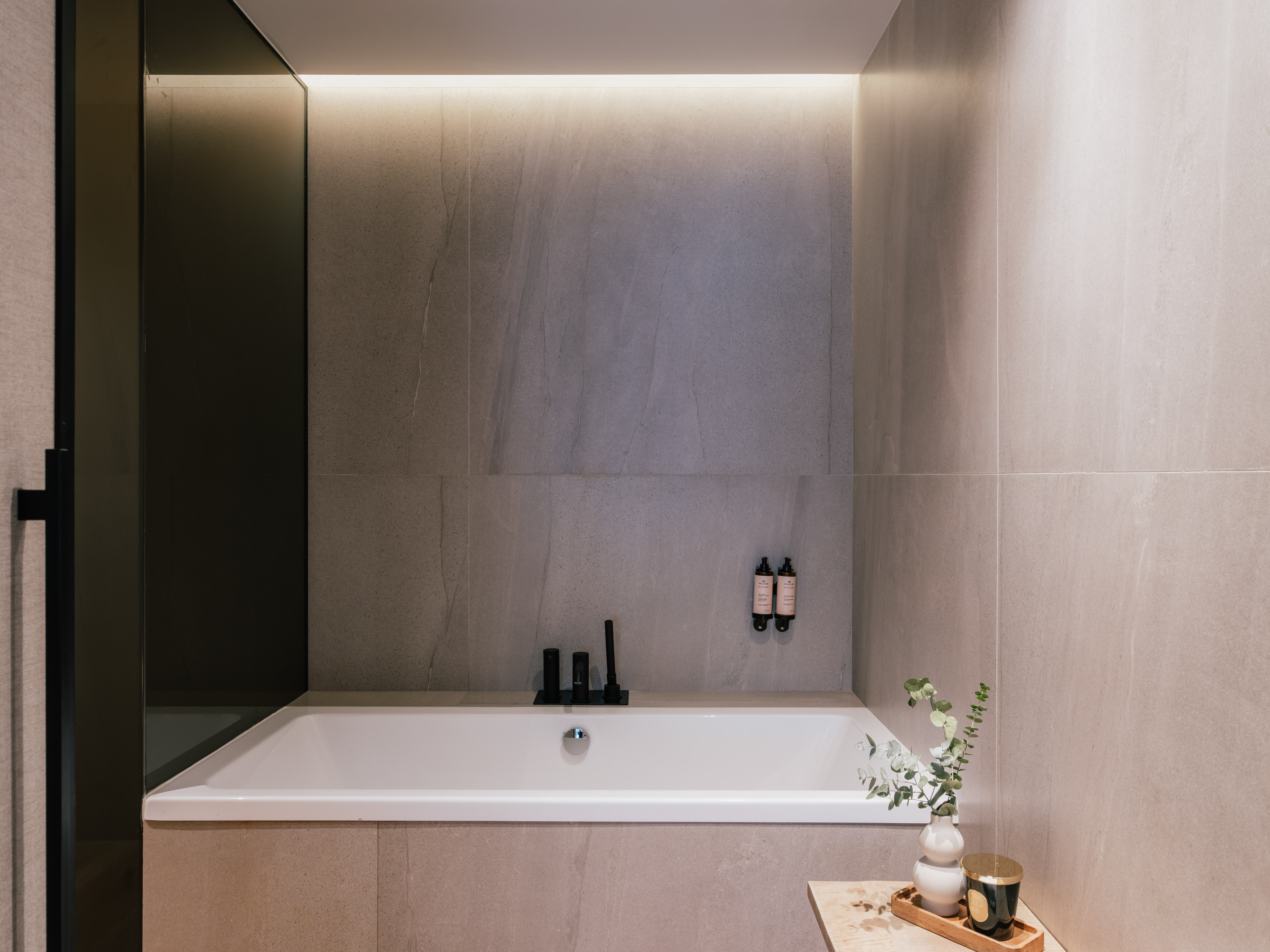 Modern bathroom with white tub, grey tiles, black fixtures, and LED lighting.