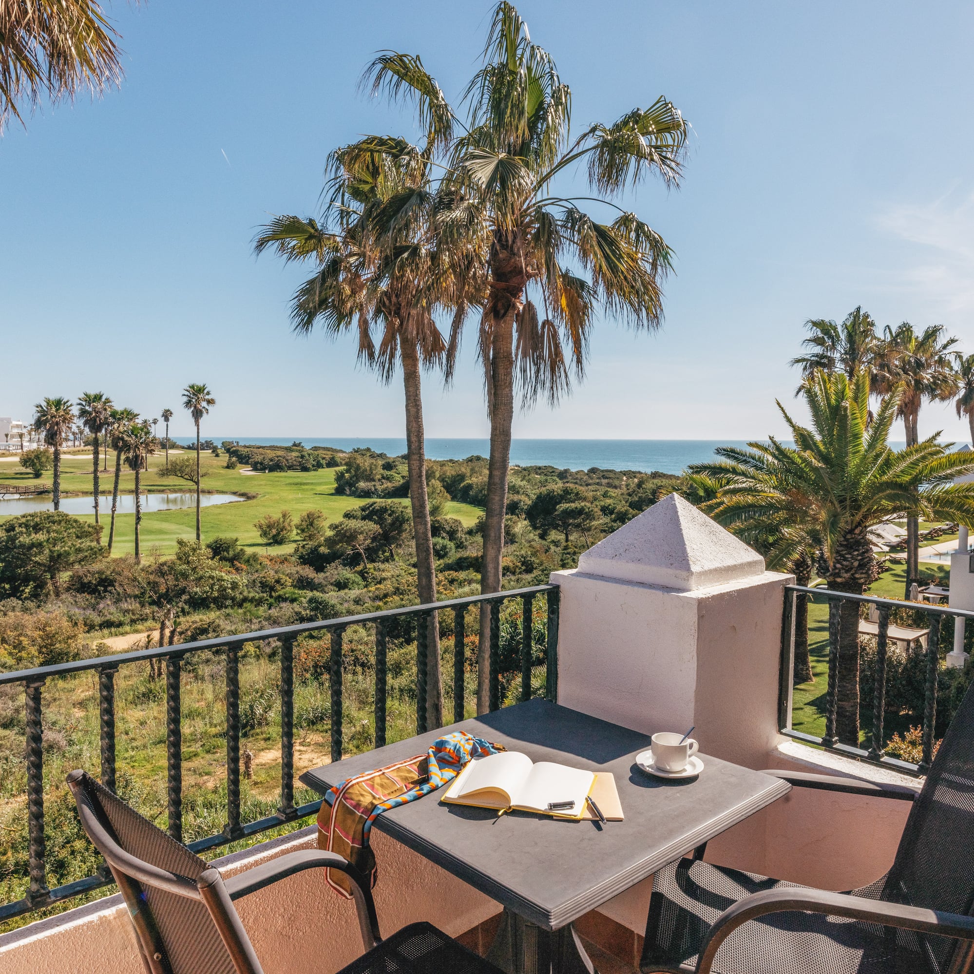 Resort balcony with golf course, ocean view, palm trees, coffee, and book.