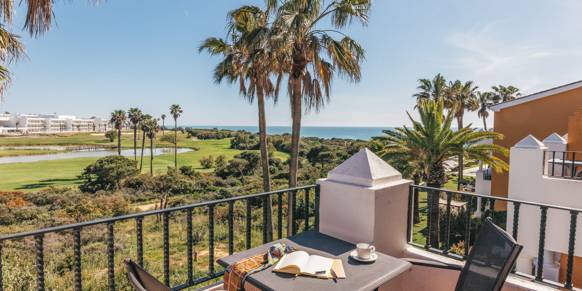 Resort balcony with golf course, ocean view, palm trees, coffee, and book.