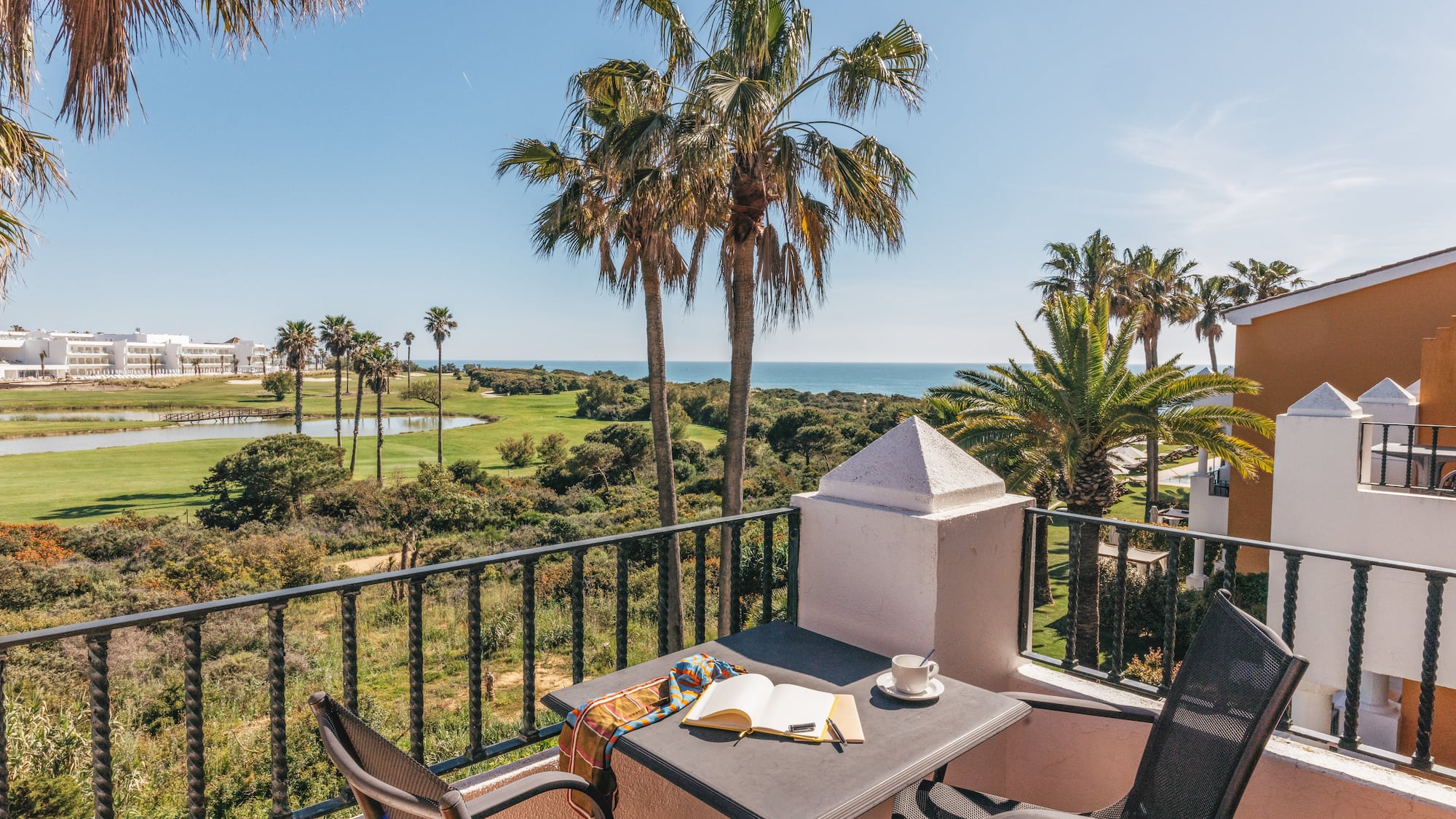 Resort balcony with golf course, ocean view, palm trees, coffee, and book.