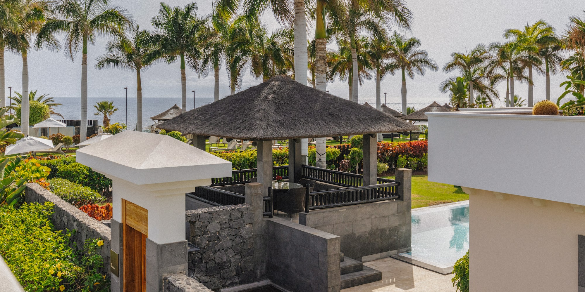 Lush tropical resort with thatched gazebo, palm trees, pool, and ocean view.