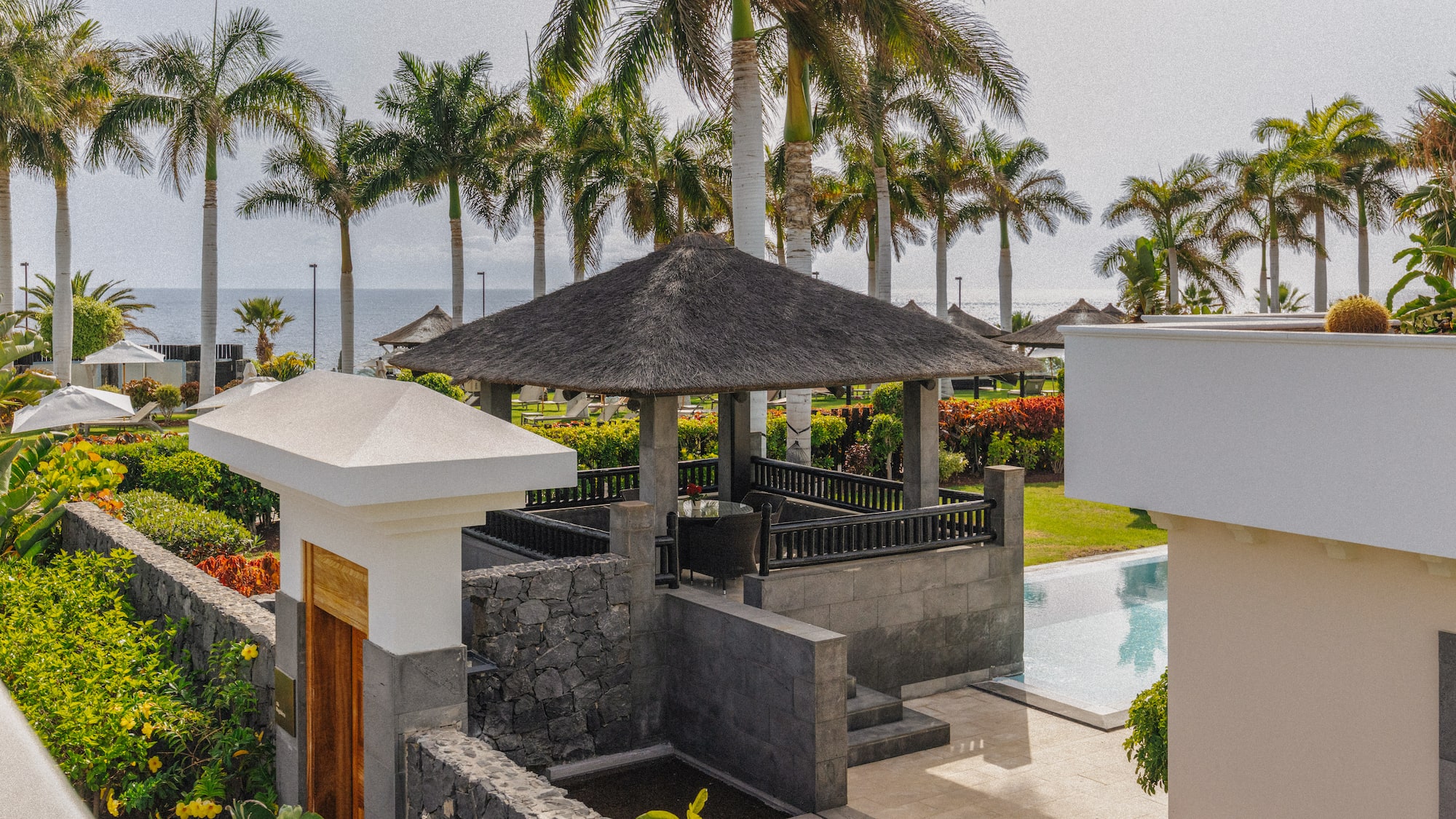Lush tropical resort with thatched gazebo, palm trees, pool, and ocean view.
