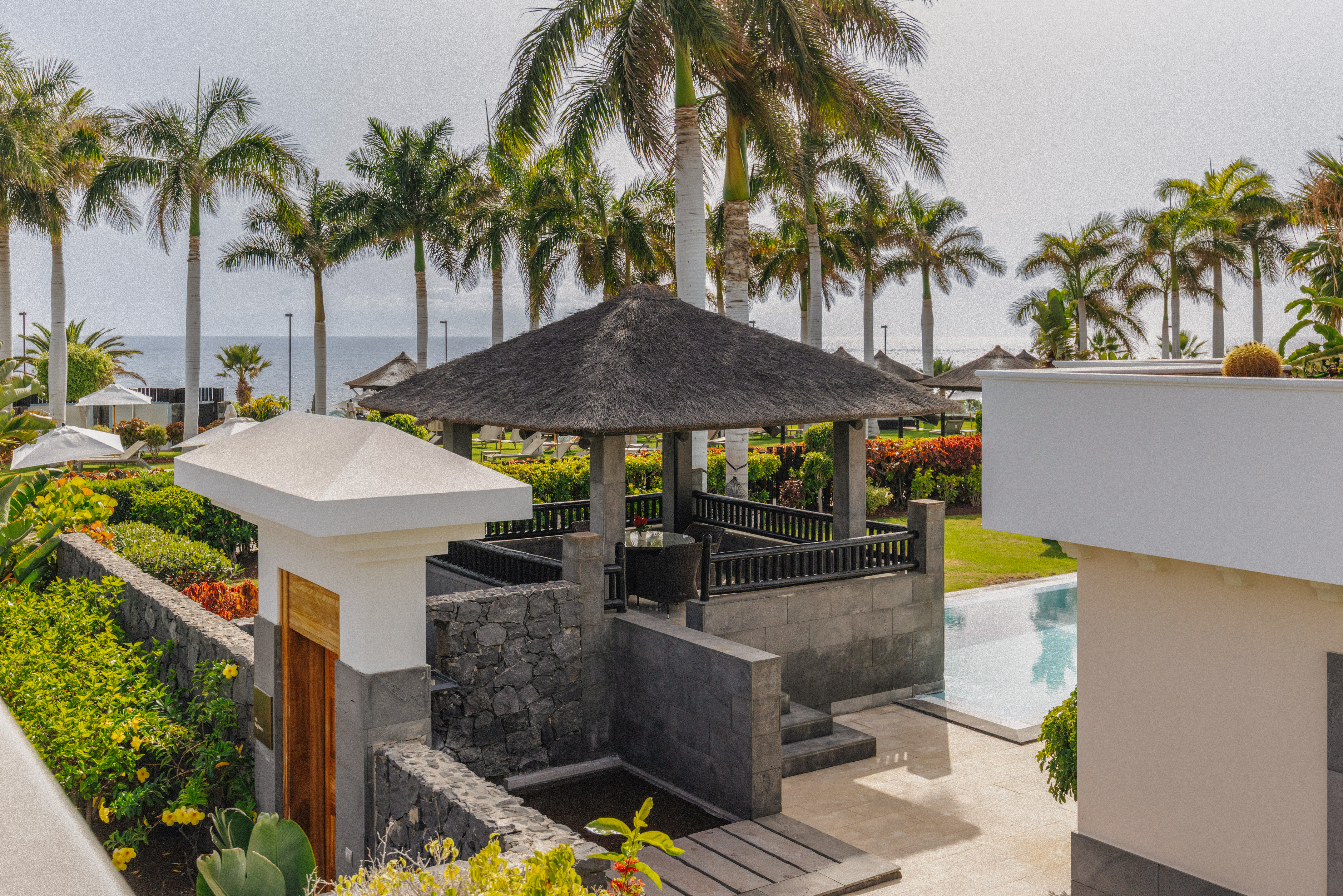 Lush tropical resort with thatched gazebo, palm trees, pool, and ocean view.