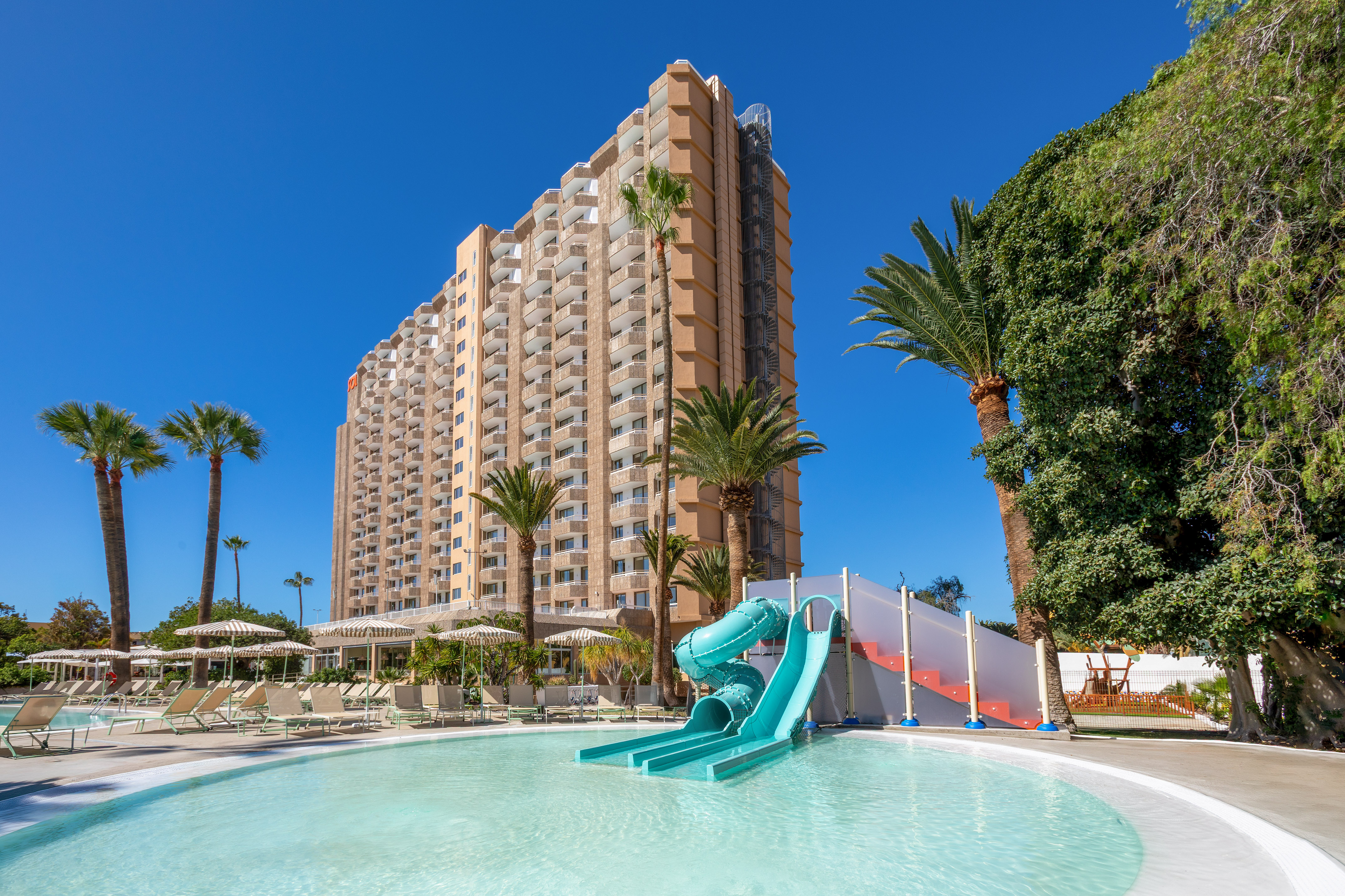 Sunny hotel resort with large pool, turquoise water slide, and tall palm trees.