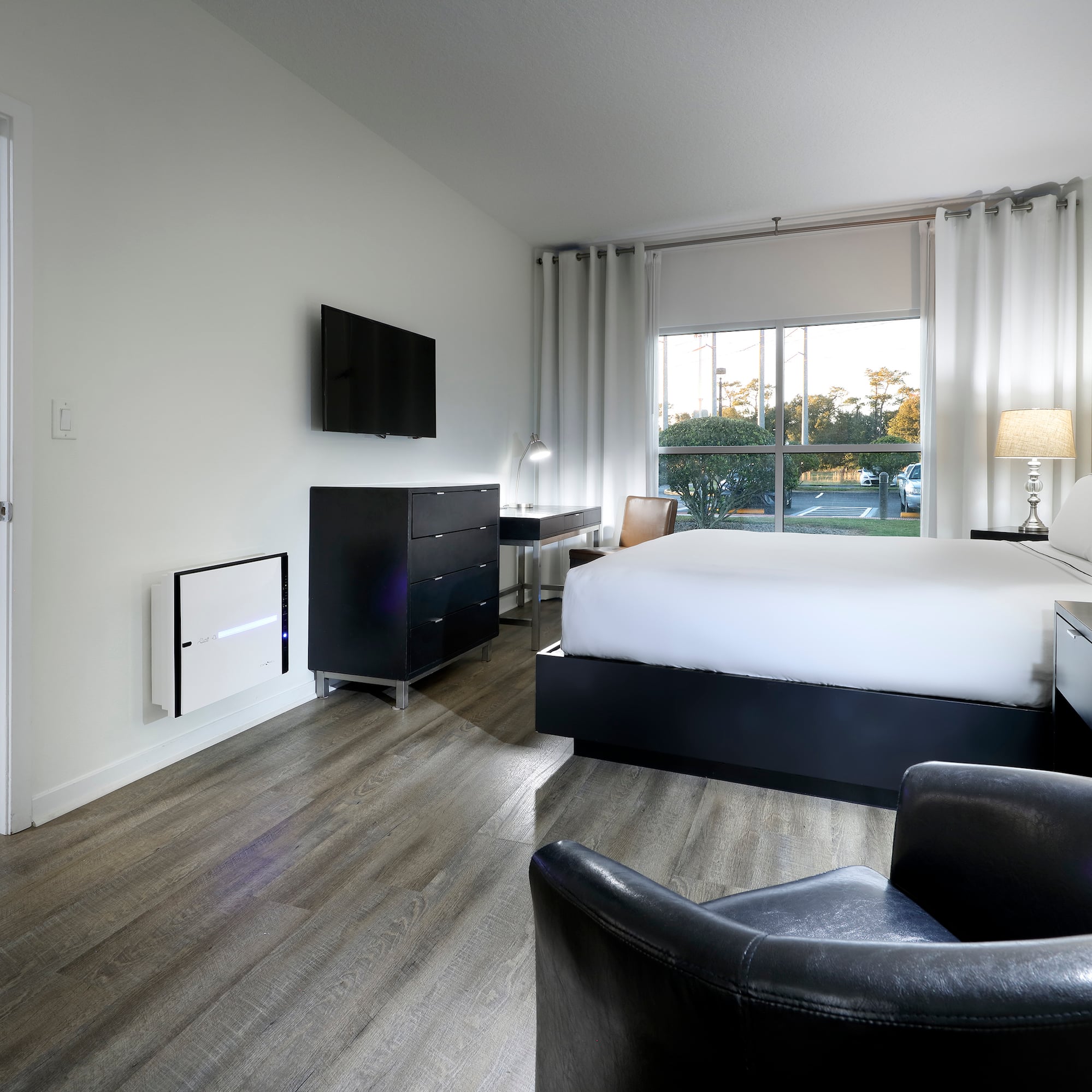 Modern hotel bedroom with bed, TV, large window, wood floor, and living area.