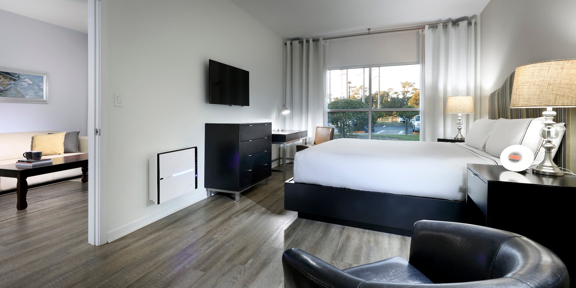 Modern hotel bedroom with bed, TV, large window, wood floor, and living area.