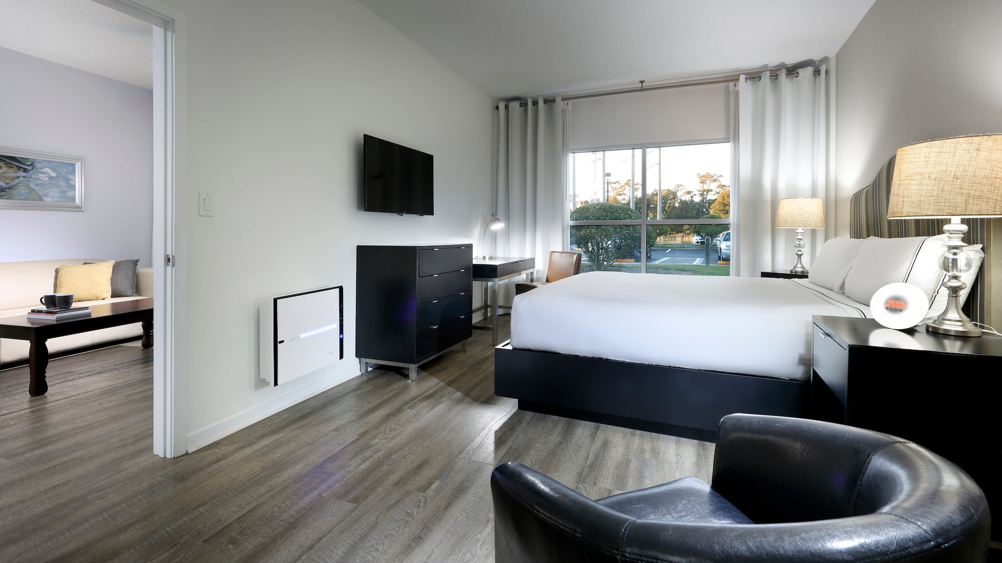 Modern hotel bedroom with bed, TV, large window, wood floor, and living area.