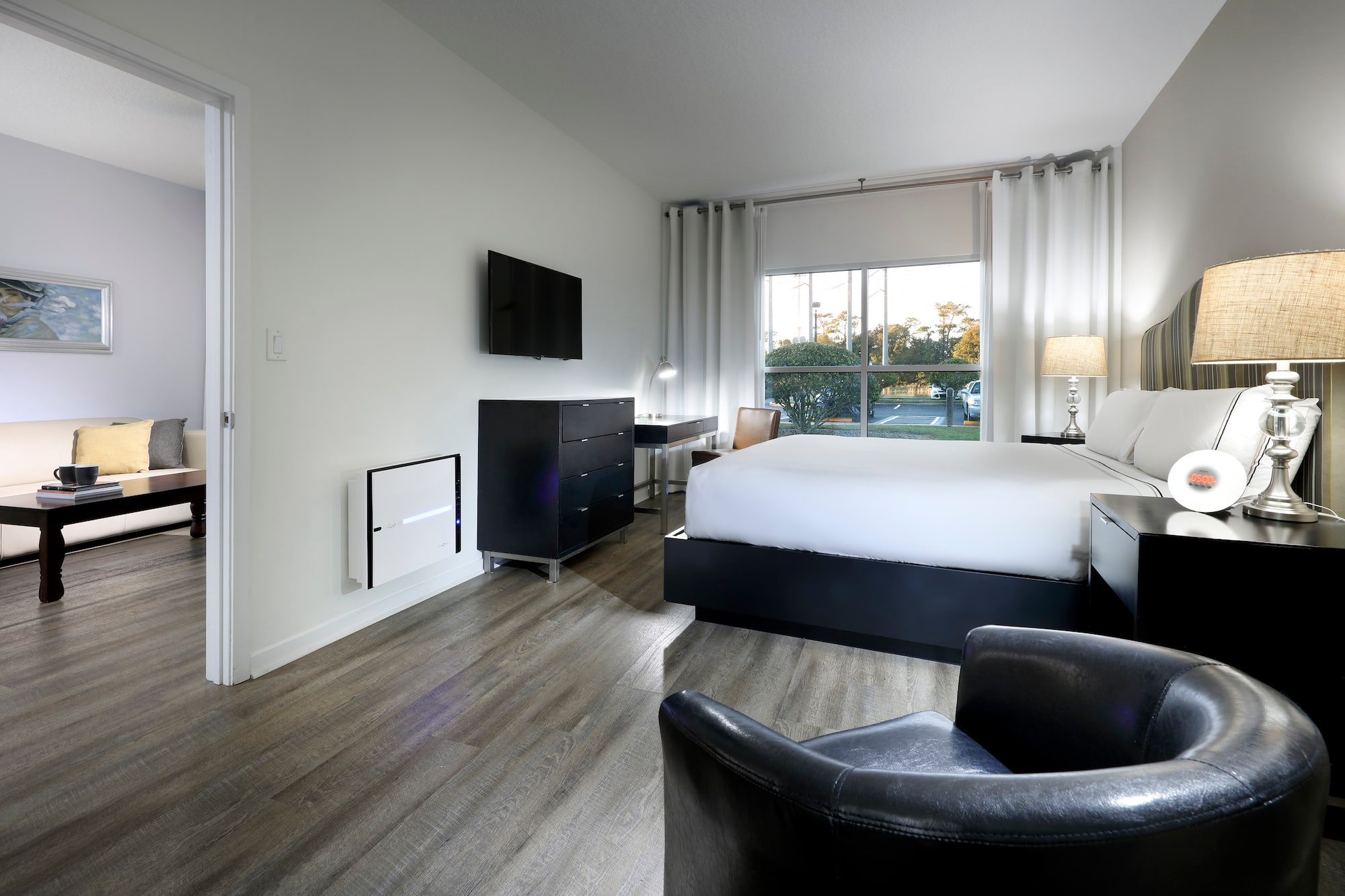 Modern hotel bedroom with bed, TV, large window, wood floor, and living area.