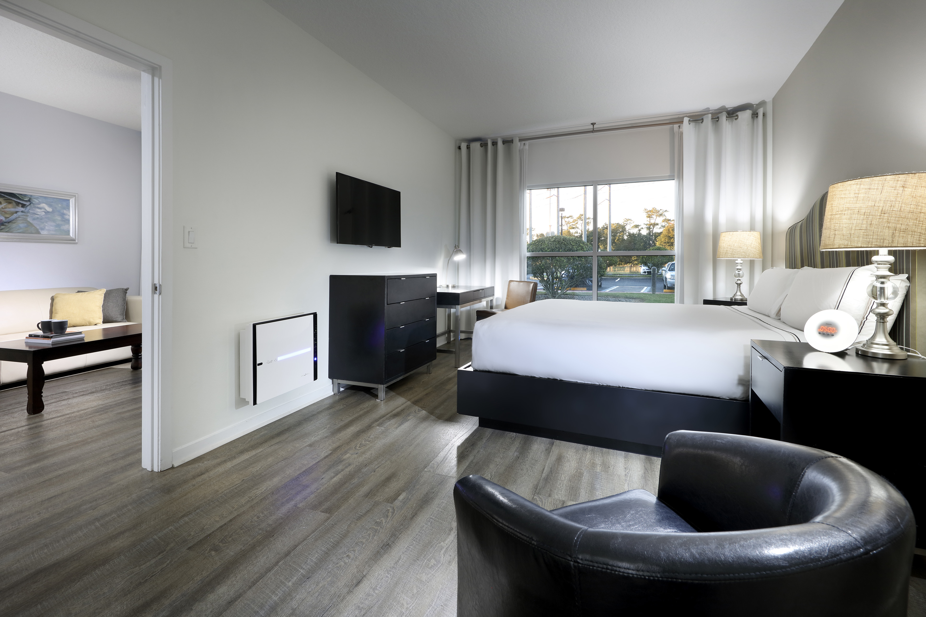 Modern hotel bedroom with bed, TV, large window, wood floor, and living area.