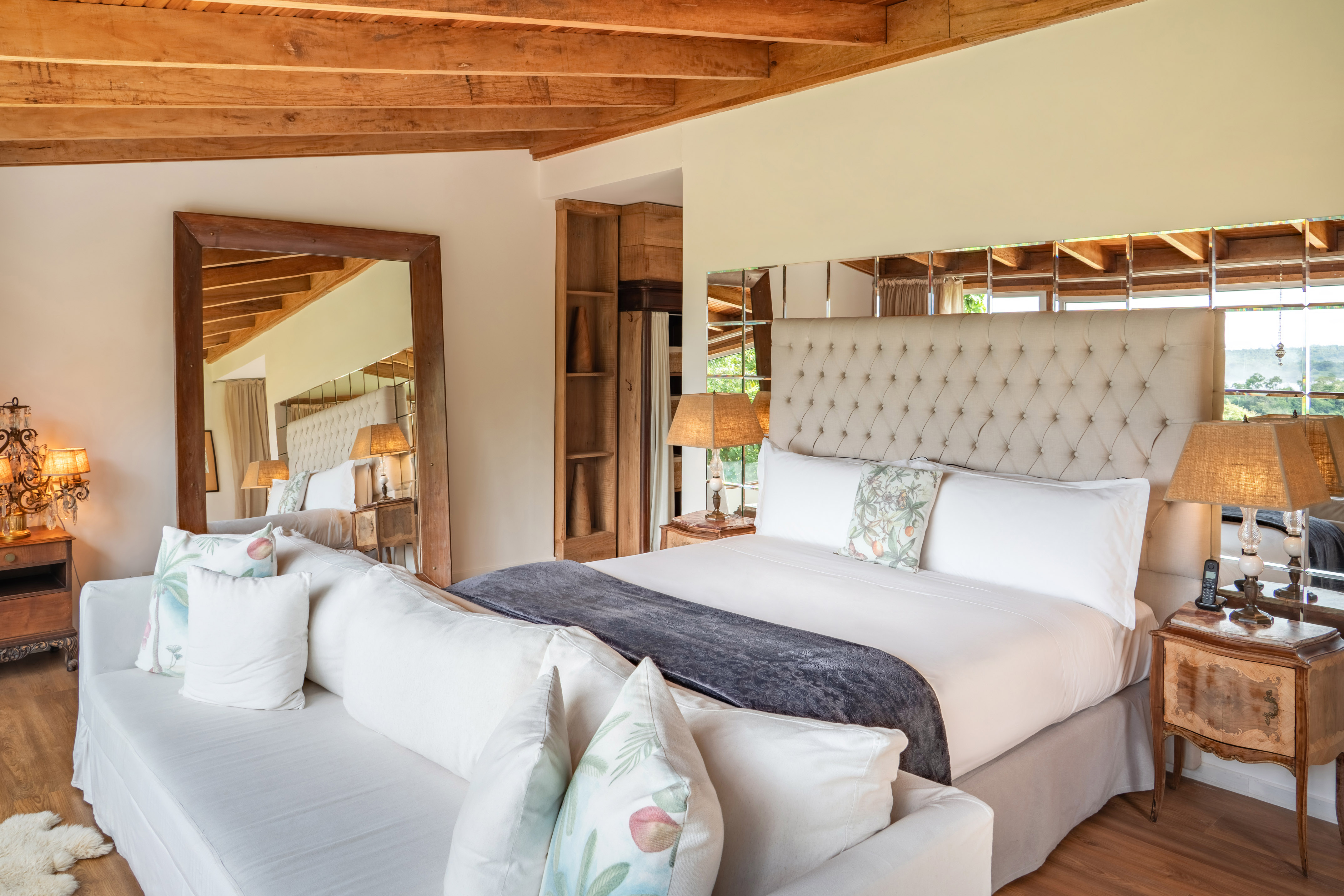 Luxury bedroom with tufted bed, wood beams, white sofa, and rustic furnishings.