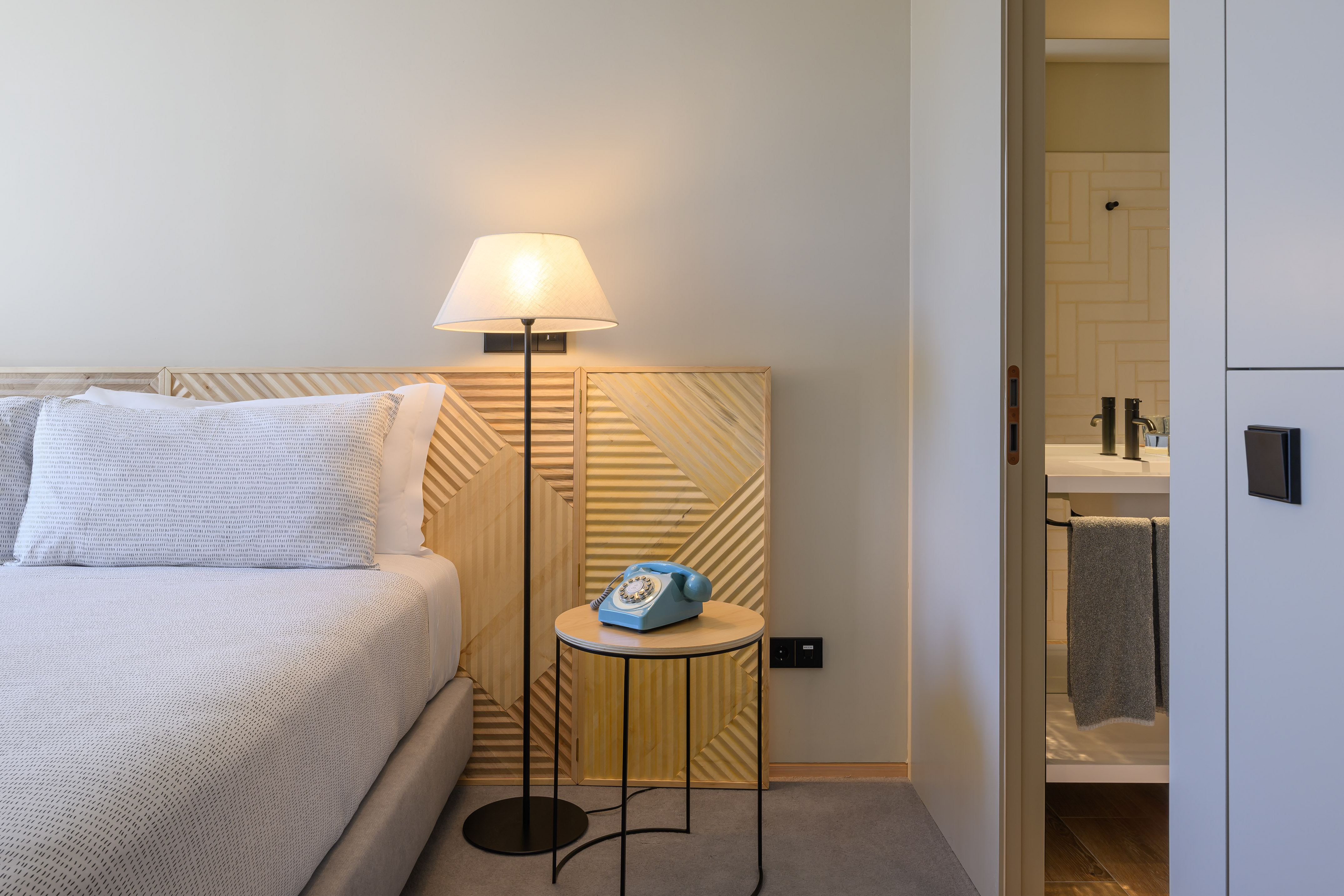 Modern hotel room with wood headboard, vintage blue phone, and bathroom entry.