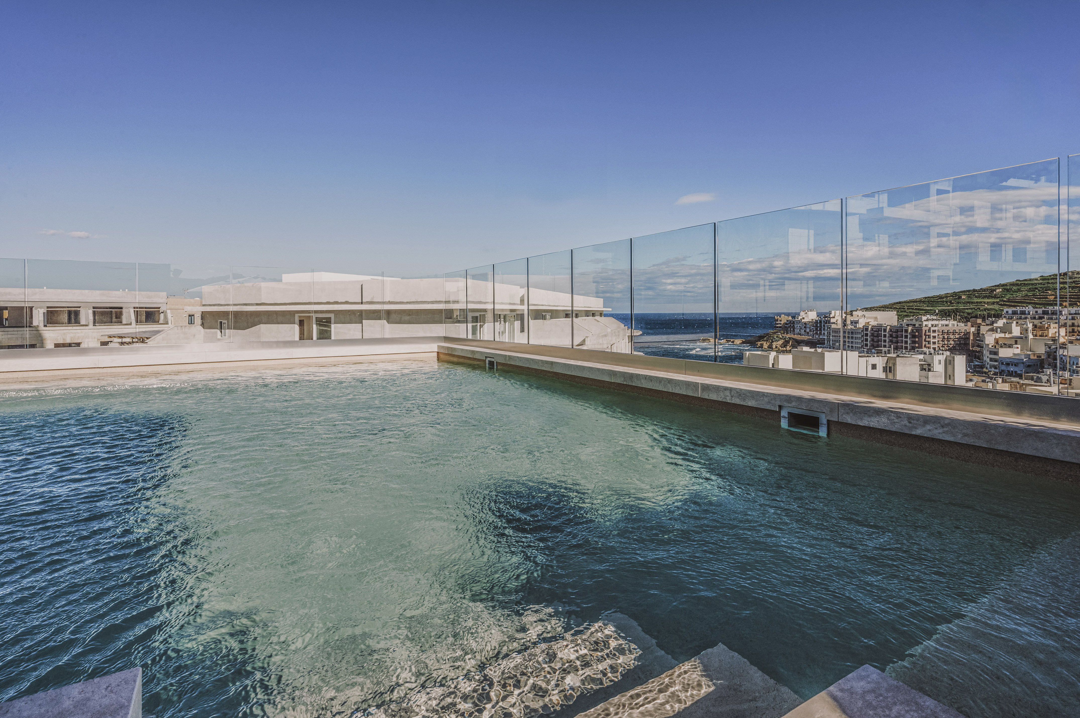 Rooftop infinity pool with glass barrier, ocean, and coastal town views.