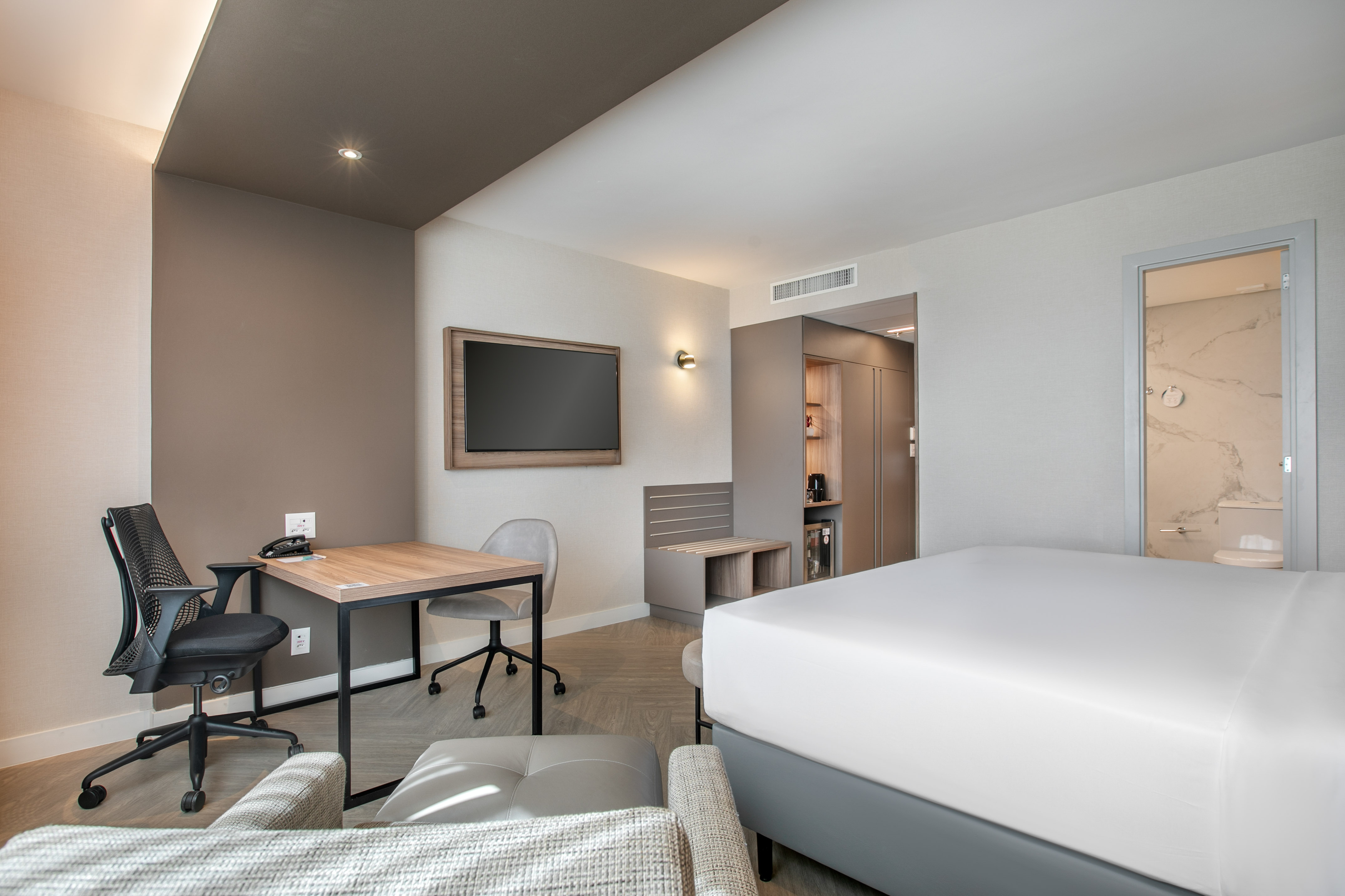 Modern hotel room with king bed, desk, wall-mounted TV, and a visible bathroom.