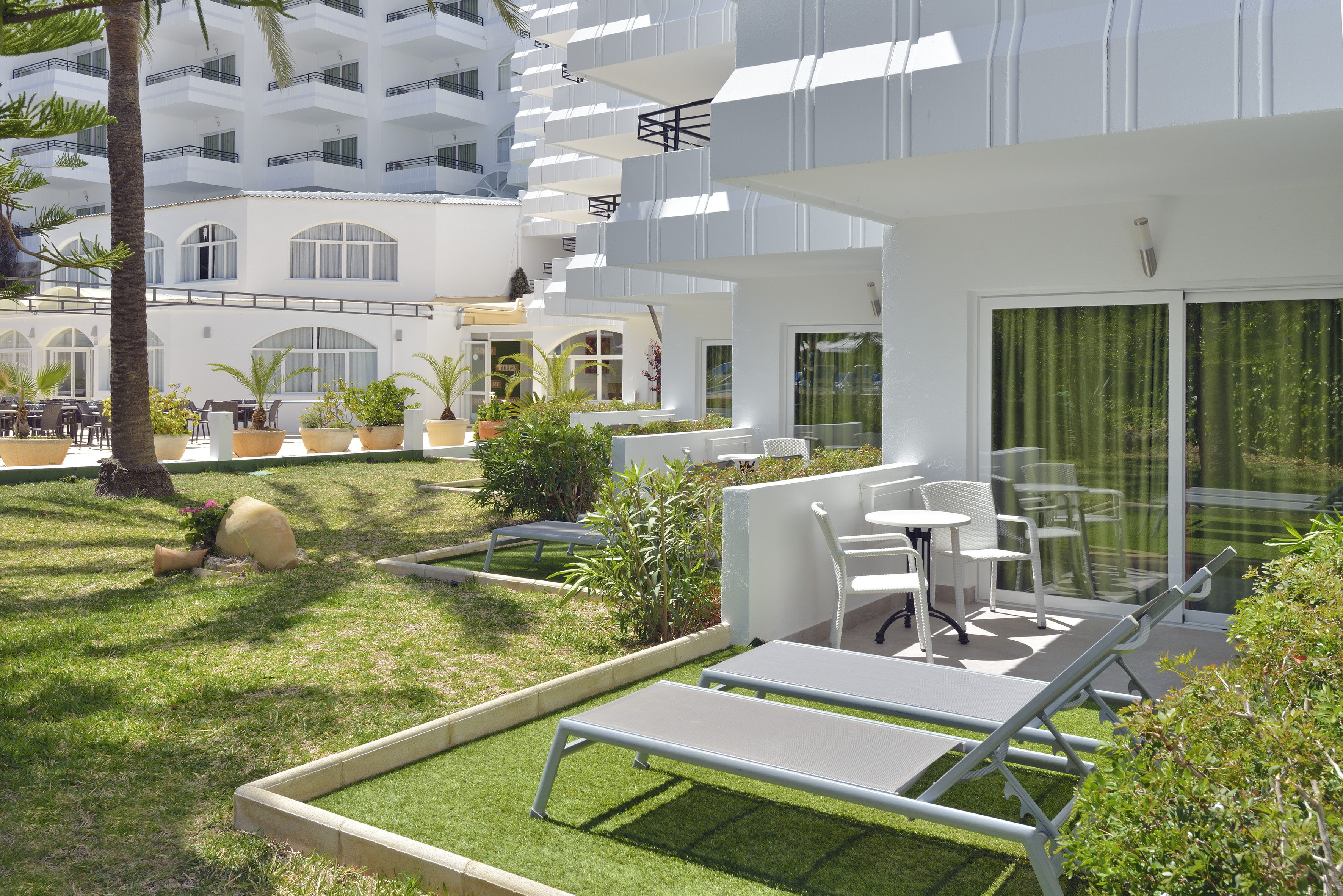 Hotel garden terrace with sun loungers, palm trees, and white building.