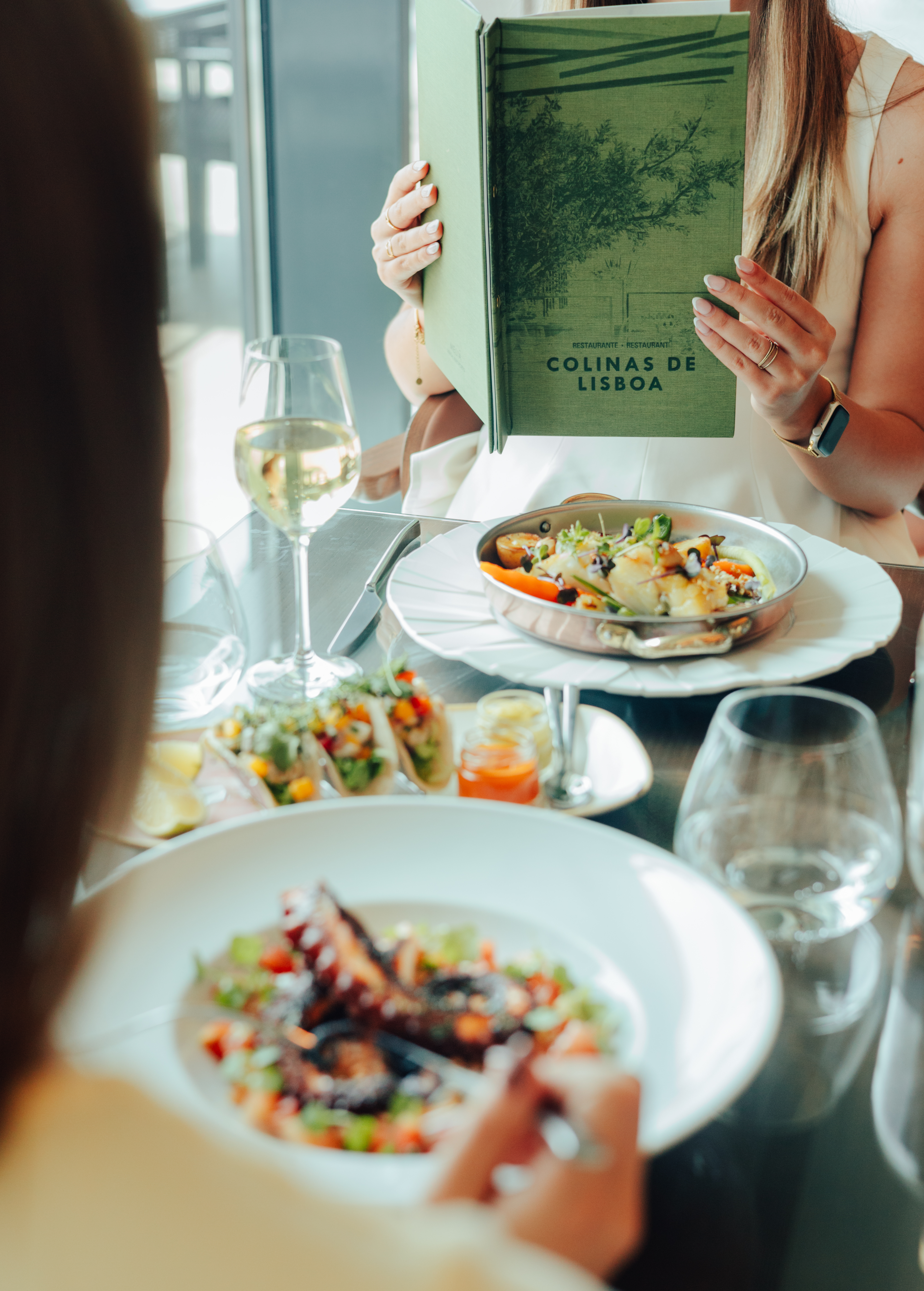 Woman holds 'Colinas de Lisboa' menu; seafood and wine on restaurant table.