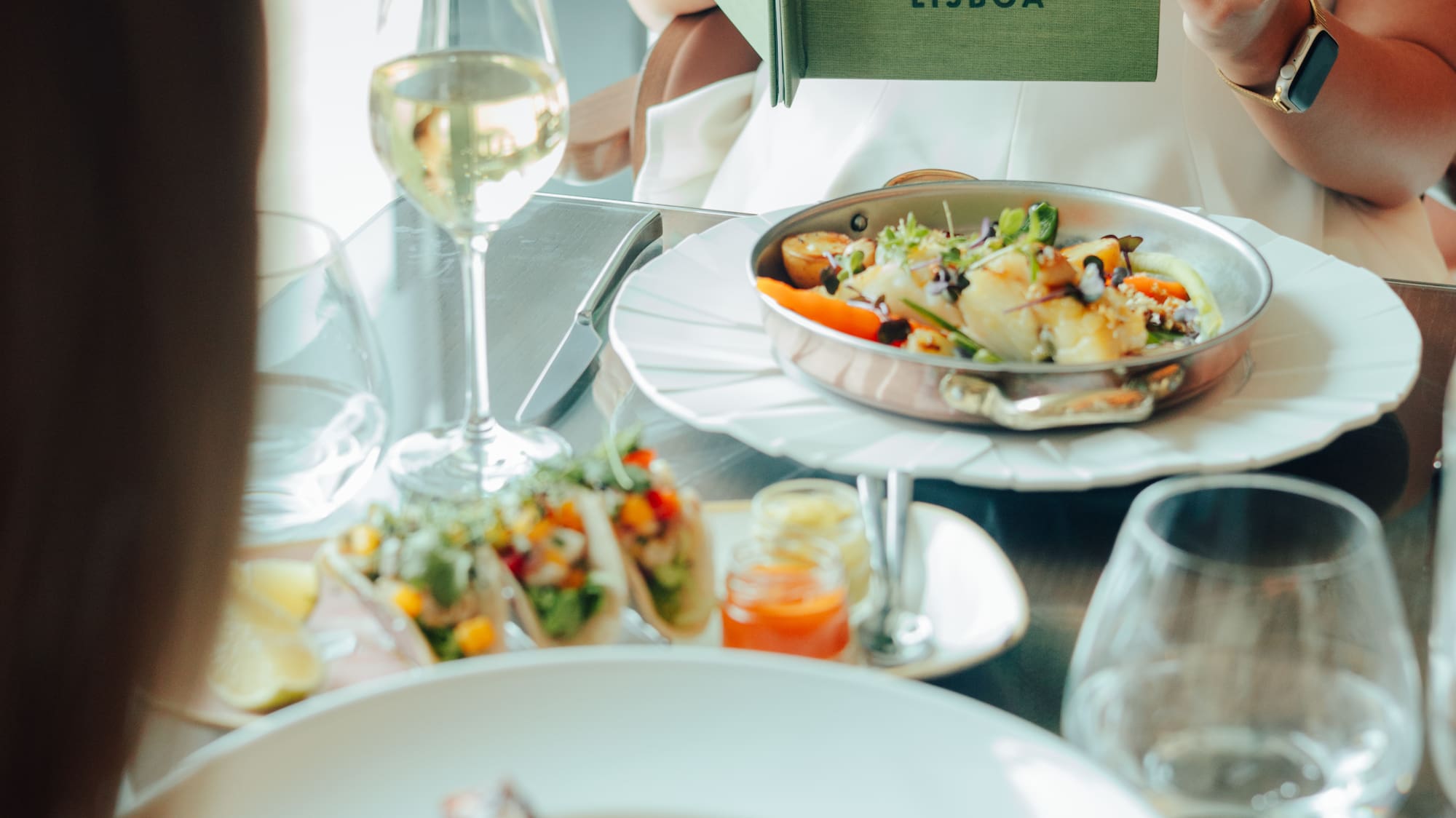 Woman holds 'Colinas de Lisboa' menu; seafood and wine on restaurant table.