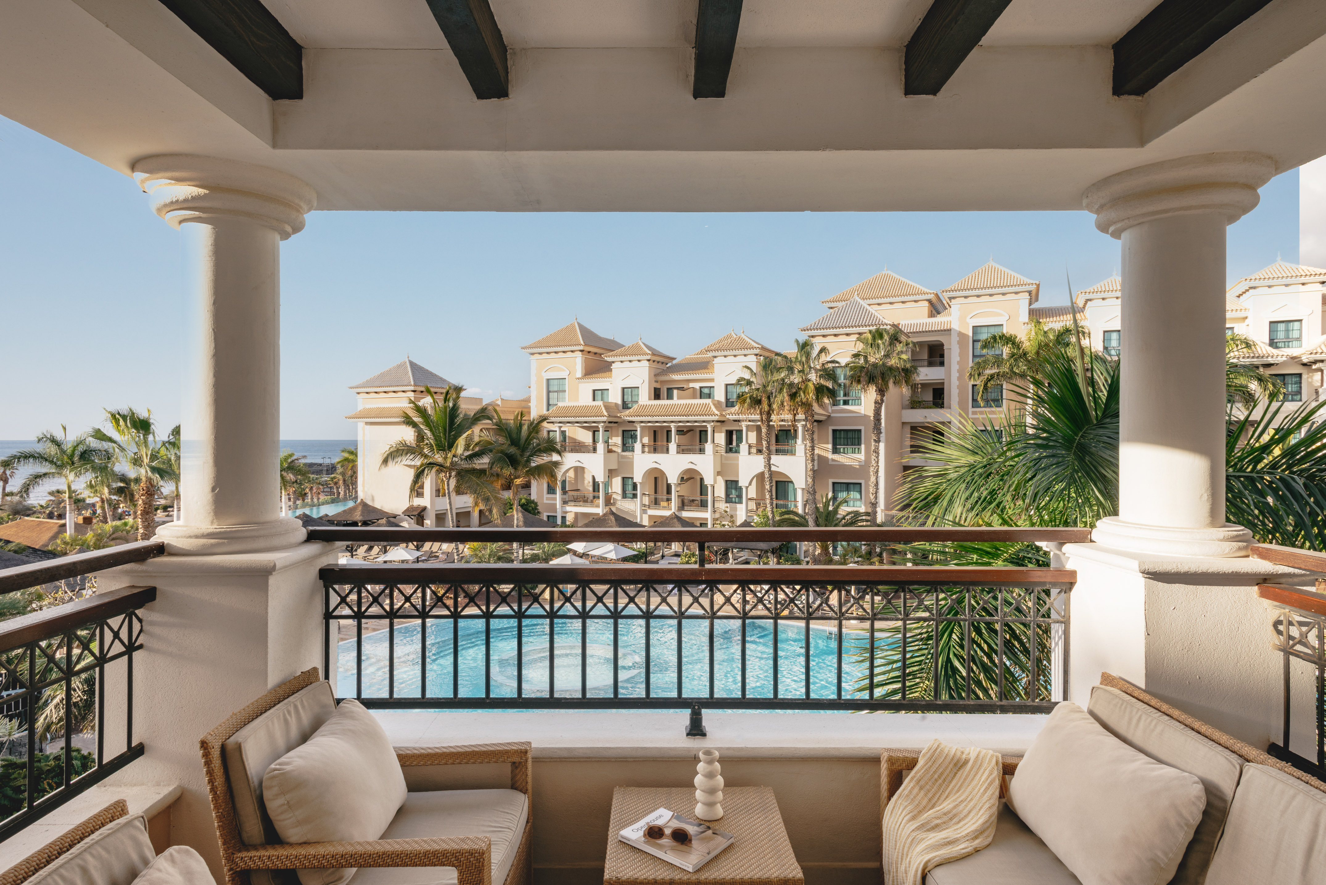 Balcony overlooking luxury resort pool, palm trees, hotel, and ocean.