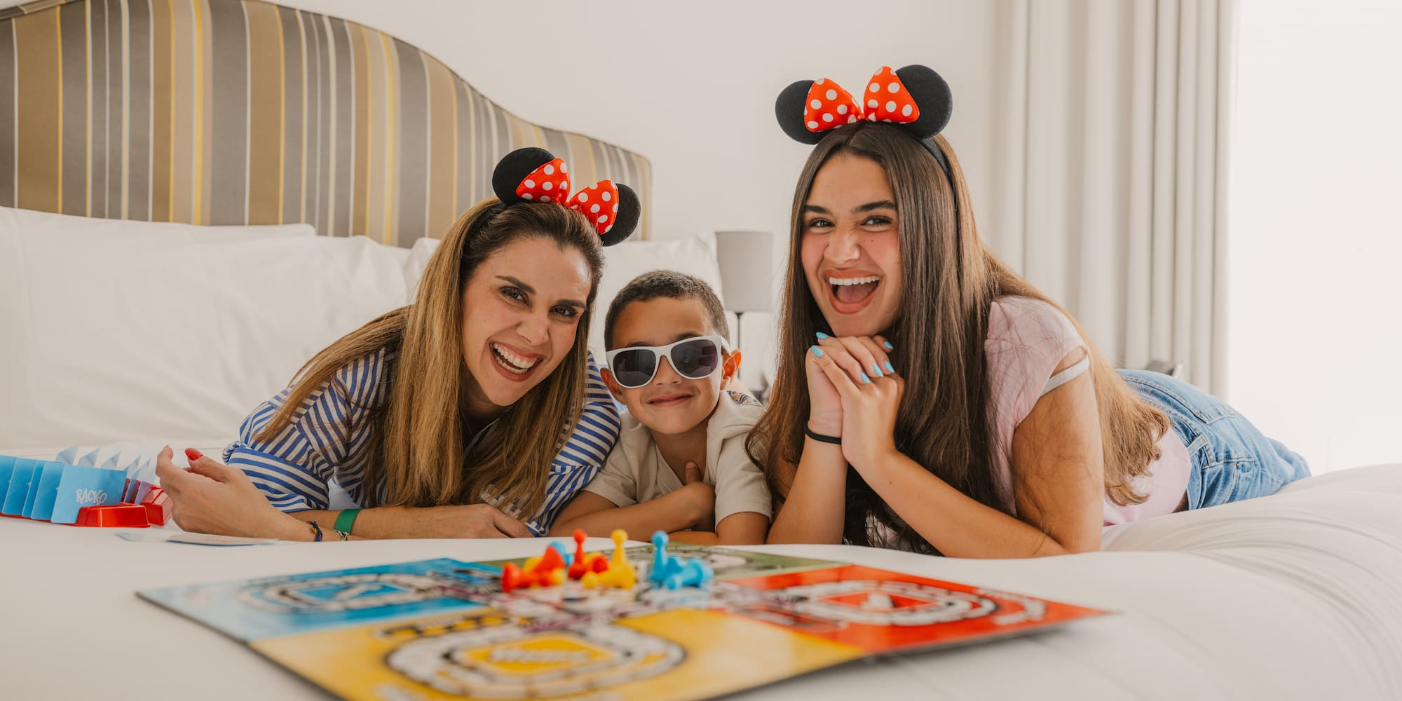 Smiling family with Minnie Mouse ears playing board game on hotel bed.
