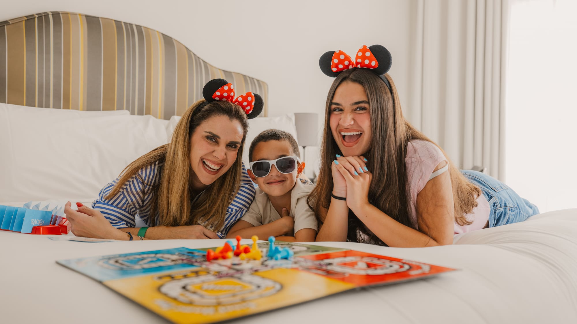 Smiling family with Minnie Mouse ears playing board game on hotel bed.