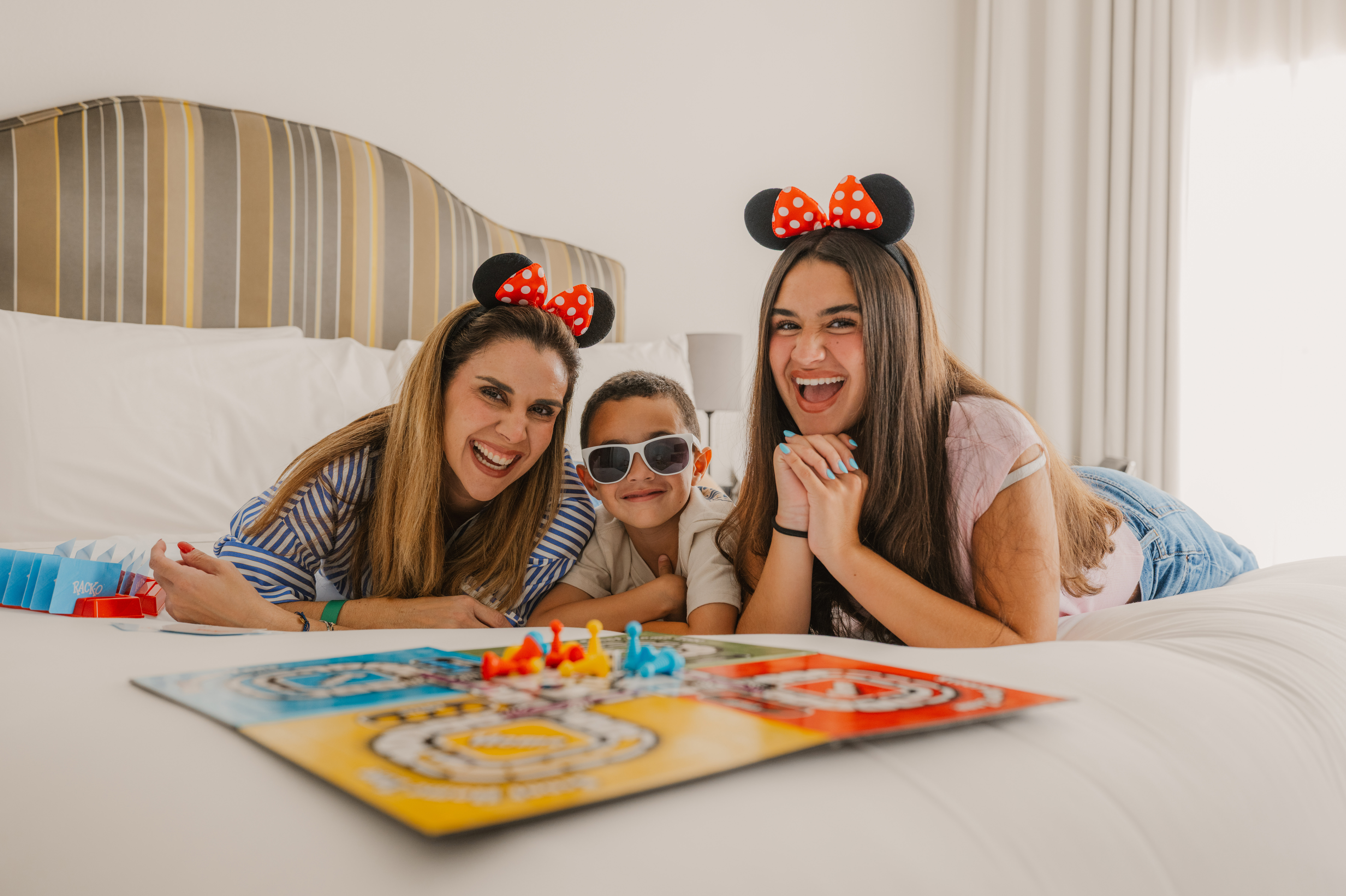 Smiling family with Minnie Mouse ears playing board game on hotel bed.