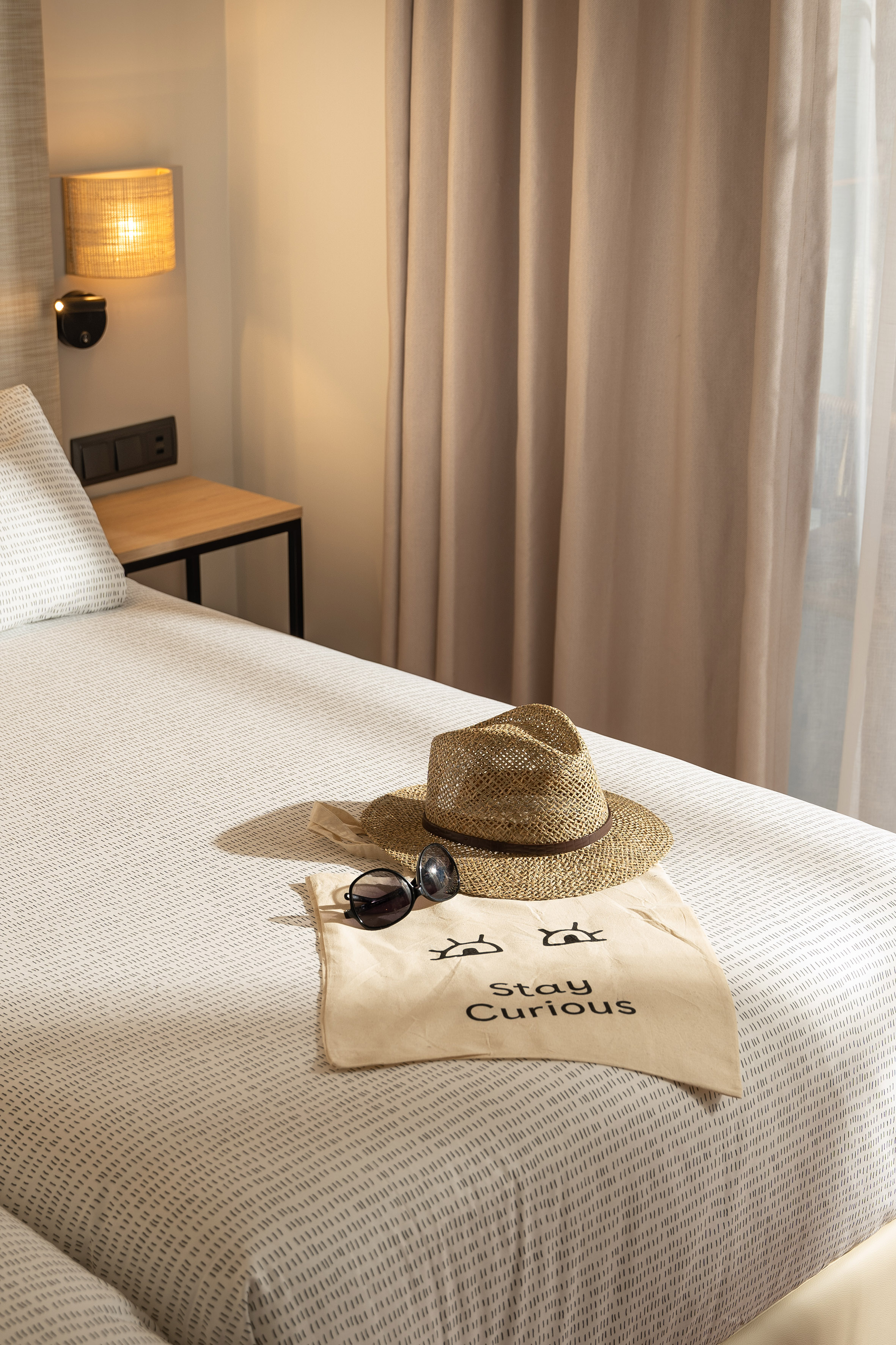 Straw hat, sunglasses, "Stay Curious" tote bag on a hotel bed.
