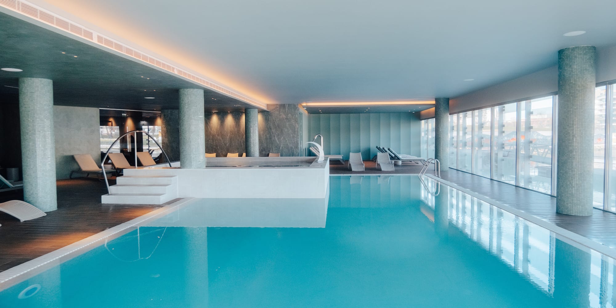 Luxury indoor swimming pool and spa with lounge chairs and large windows.