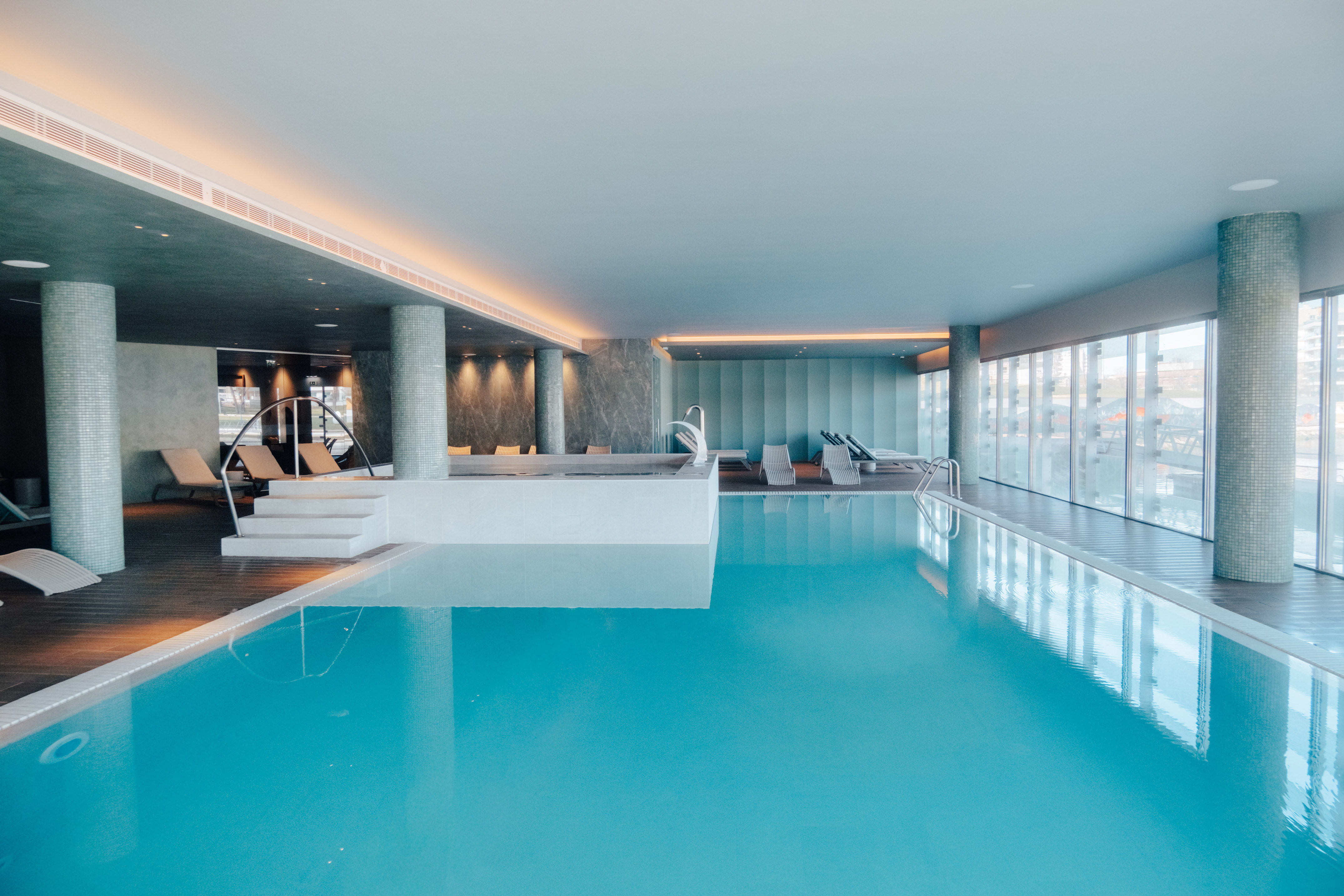 Modern indoor swimming pool with lounge chairs, large windows, and natural light.