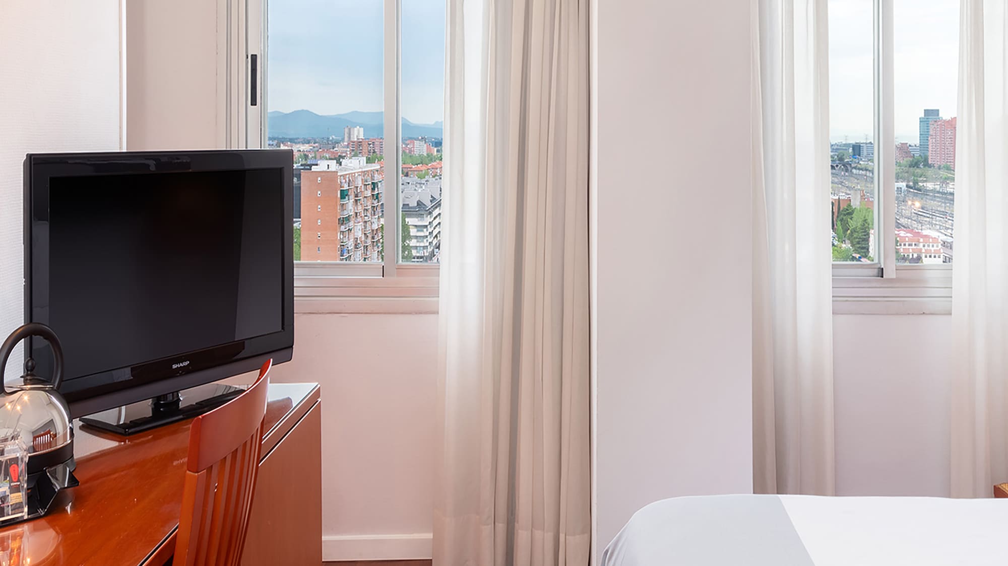 Bright hotel room featuring desk, TV, bed, and urban window view.