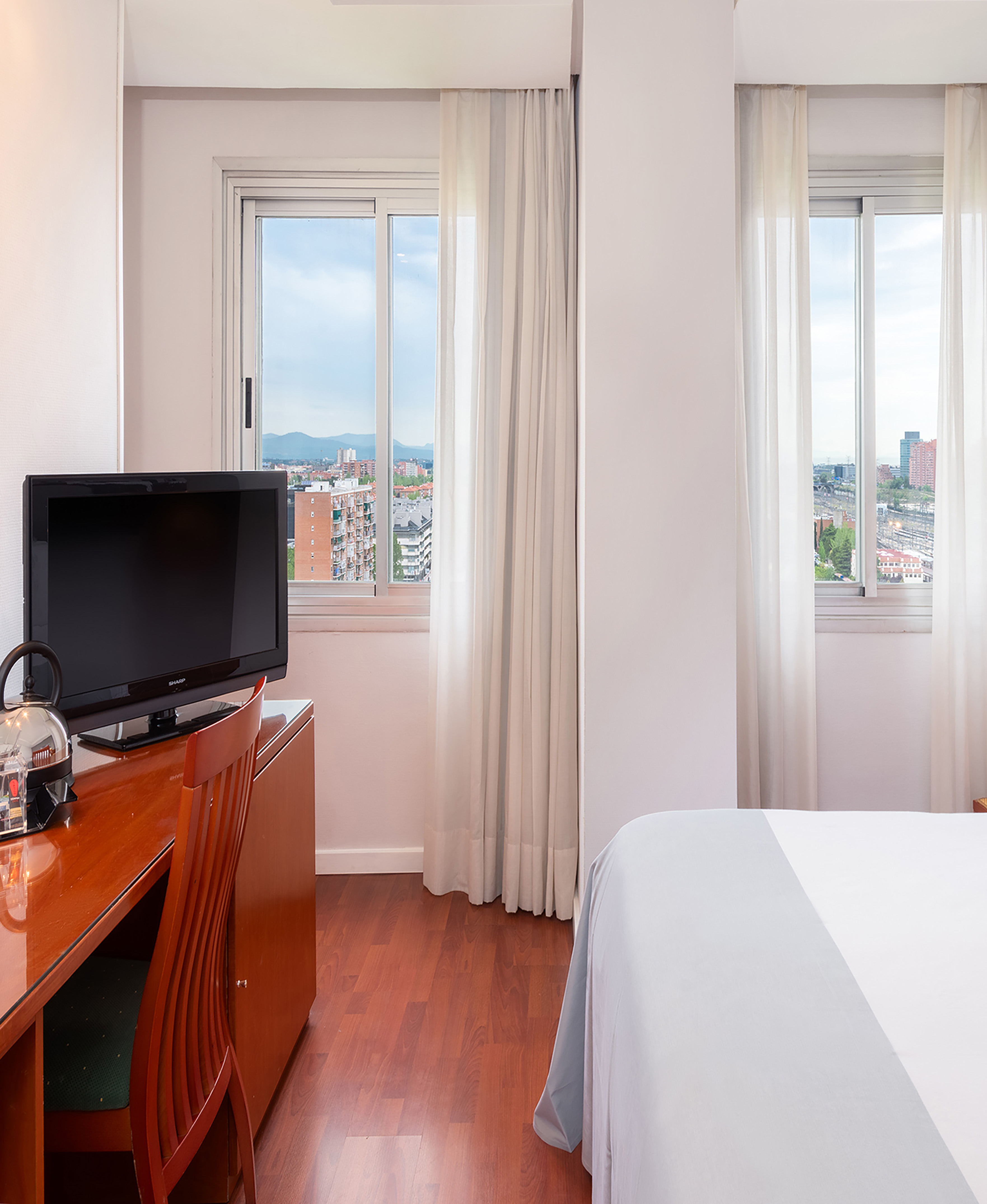 Bright hotel room featuring desk, TV, bed, and urban window view.