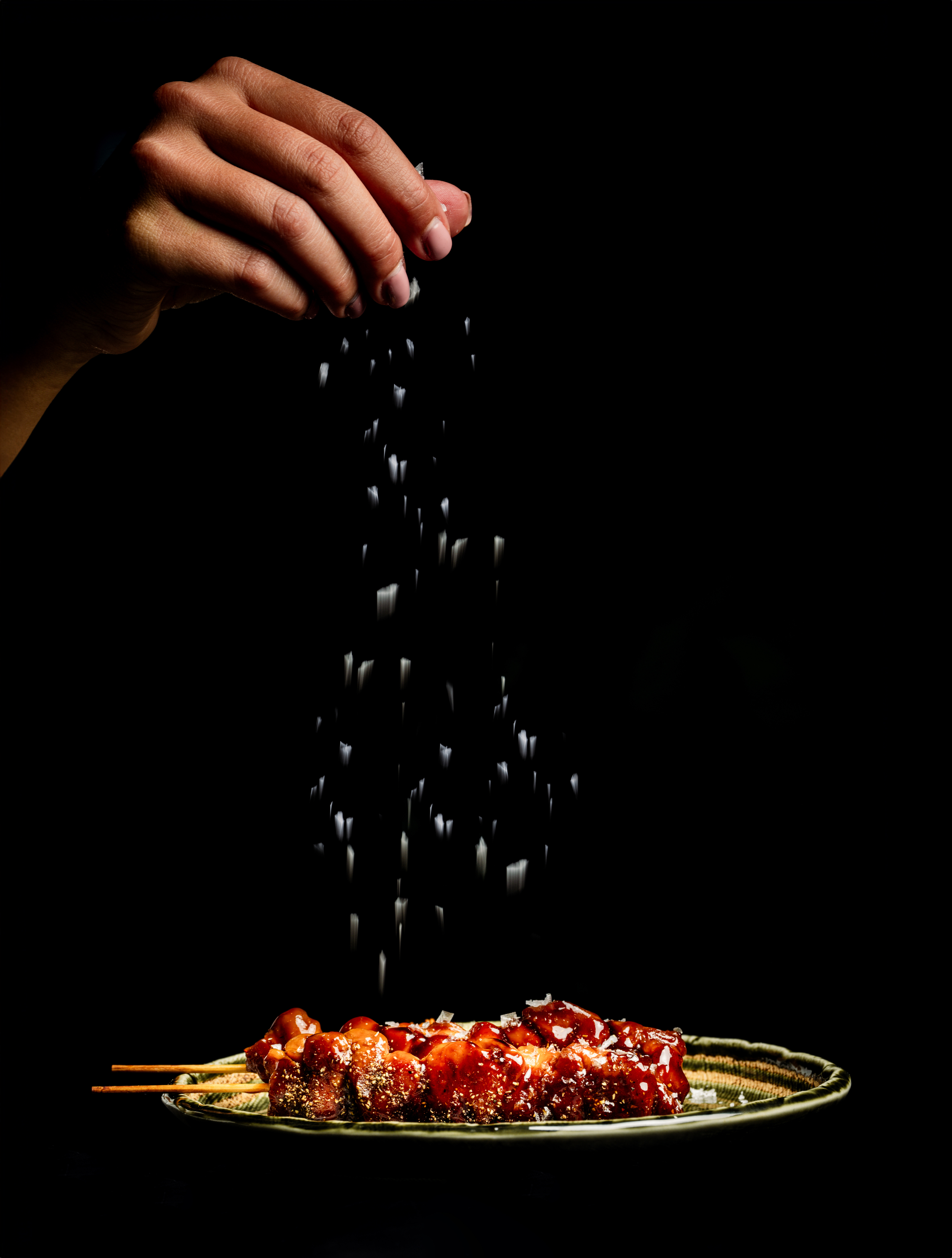Hand sprinkling salt on glazed yakitori skewers against a dark background.