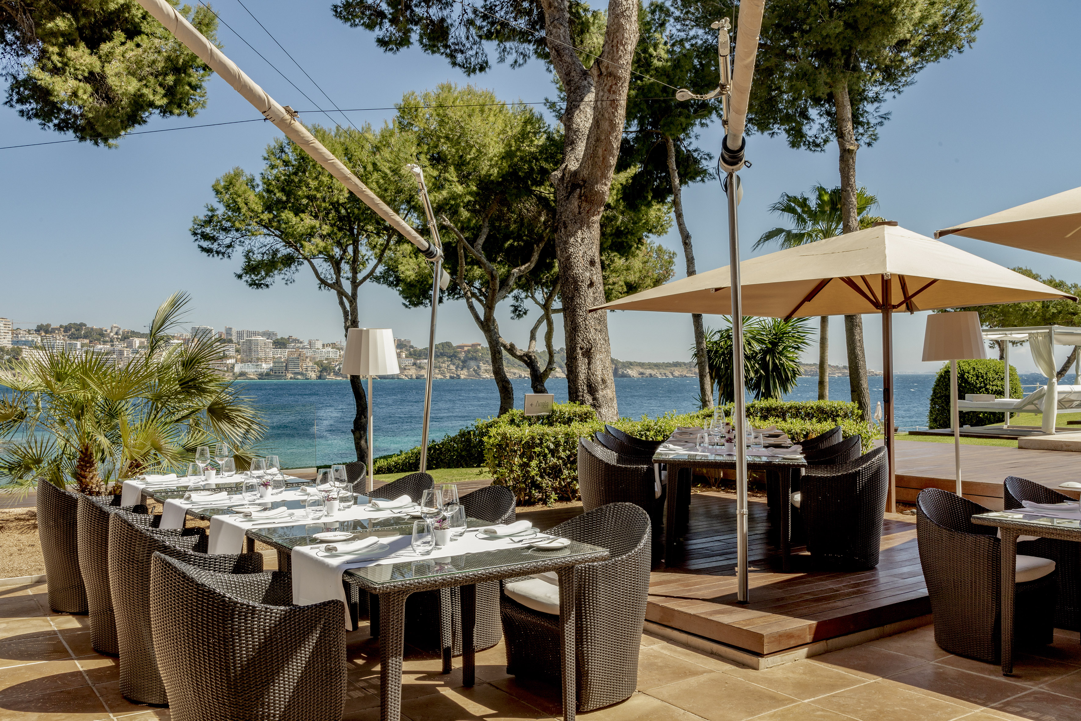 Outdoor dining terrace with set tables, blue sea view, and lush trees.