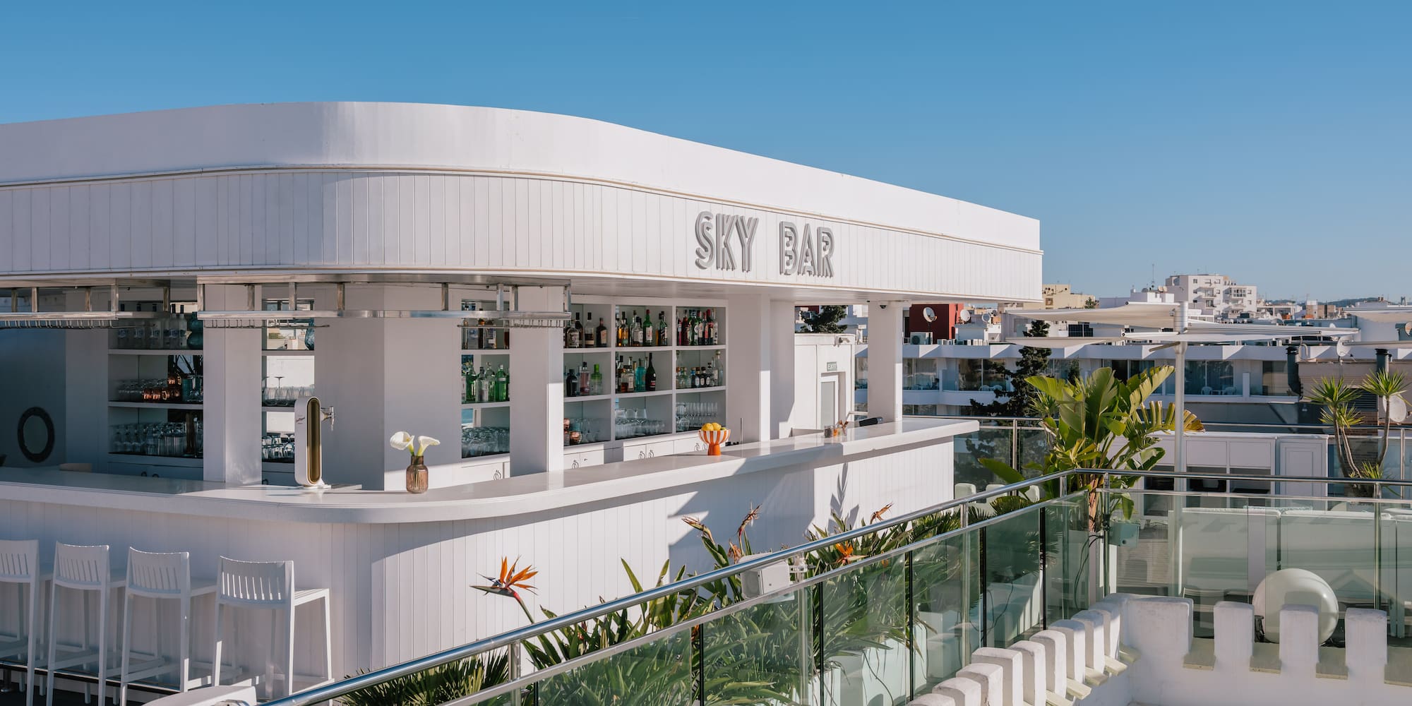 White rooftop Sky Bar with modern design, drinks, plants, and clear blue sky.