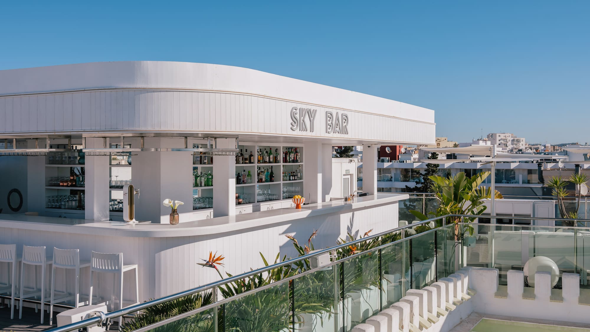 White rooftop Sky Bar with modern design, drinks, plants, and clear blue sky.