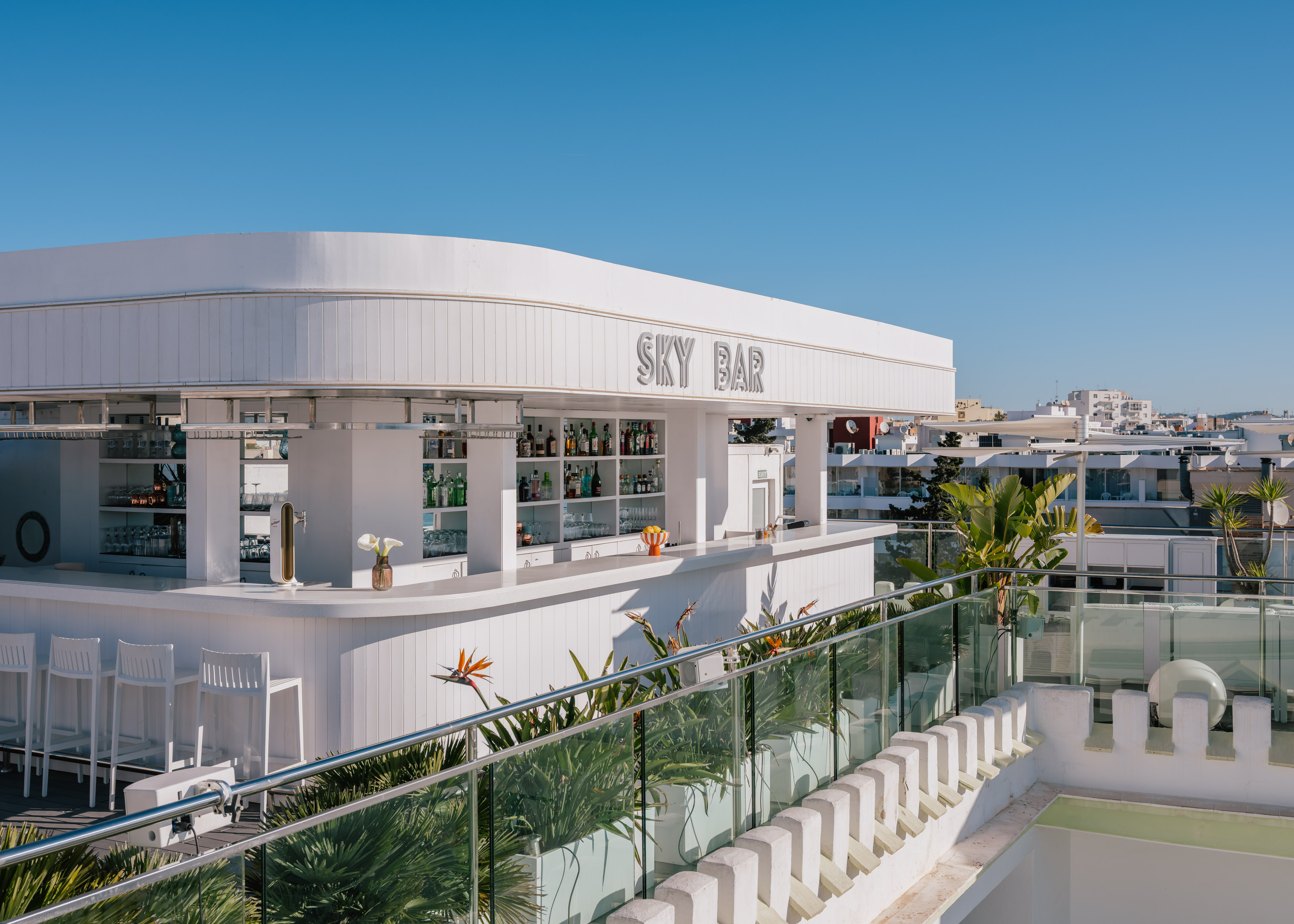 White rooftop Sky Bar with modern design, drinks, plants, and clear blue sky.